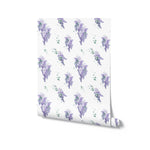 A roll of Wisteria Garden Wallpaper, with the design featuring repeating clusters of purple wisteria droplets and green leaves, suggesting an immediate connection to nature and springtime beauty.