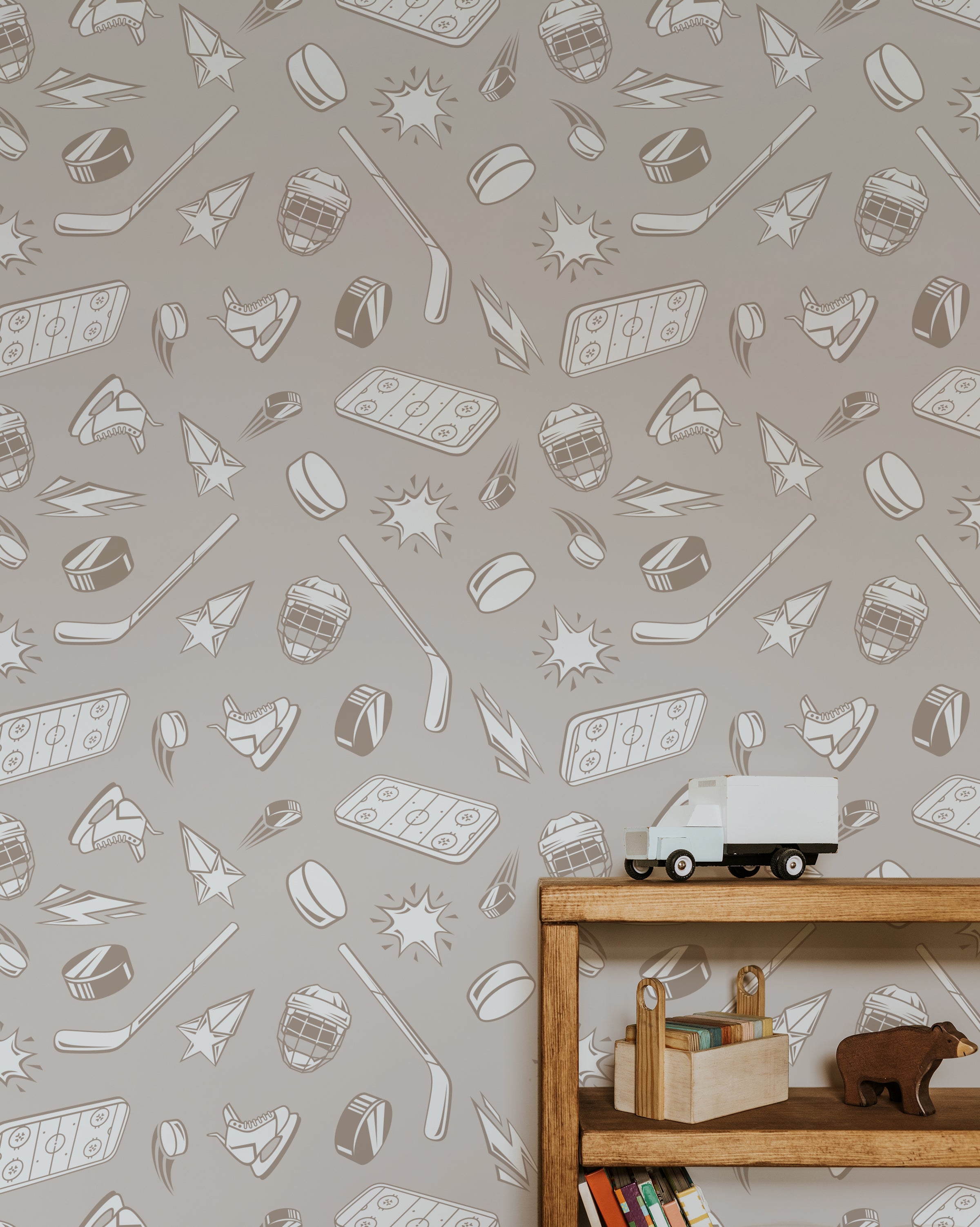 A wooden bookshelf with books and toys set against a wall covered in Ice Legend Wallpaper. The wallpaper features a playful pattern of hockey sticks, pucks, helmets, skates, and stars in beige tones.