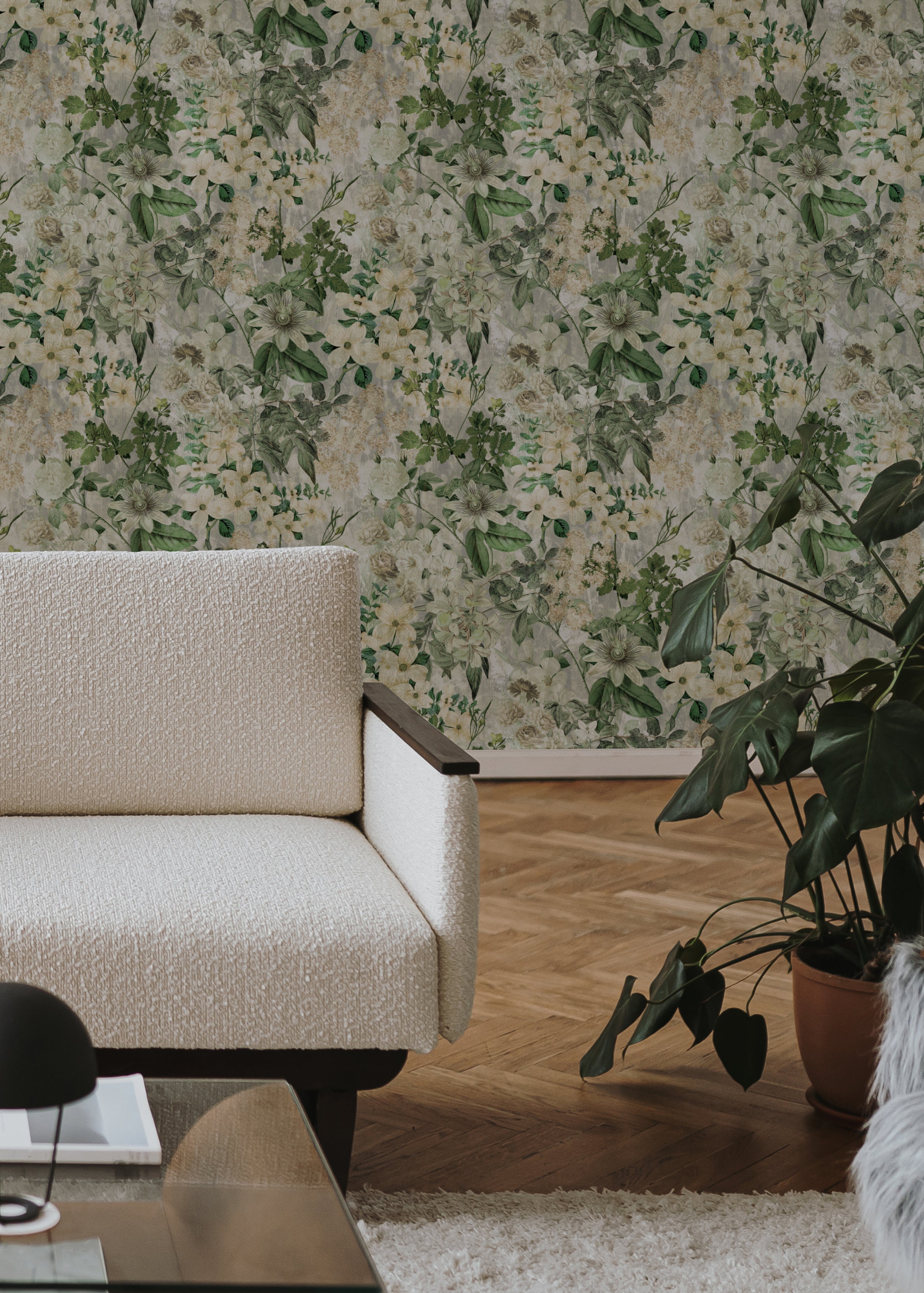 A modern living room with a white couch and indoor plants, set against a wall adorned with Floral Haven Wallpaper. The wallpaper displays an elegant floral design with green foliage and white blooms.