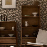 A sophisticated living space showcasing the "Daisy Blooms Wallpaper - 25"" on the walls, complemented by a wooden shelving unit displaying various pottery pieces. The naturalistic floral print enhances the room's rustic charm and creates a calming, elegant environment.