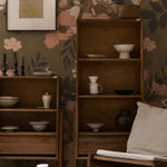 An interior scene featuring the Botanic Garden Wallpaper - 75" as a decorative backdrop in a cozy room setting. The wallpaper's large floral print in soft peach and muted green tones complements the wooden furniture and ceramic decor, enhancing the space's warm and inviting atmosphere.