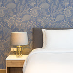 A cozy bedroom setup with a gray upholstered headboard and white bedding. A nightstand with a modern lamp sits beside the bed. The background features the Coastal Treasures Wallpaper with its detailed illustrations of seashells, coral, and sea plants in beige on a blue background, creating a serene and stylish atmosphere.