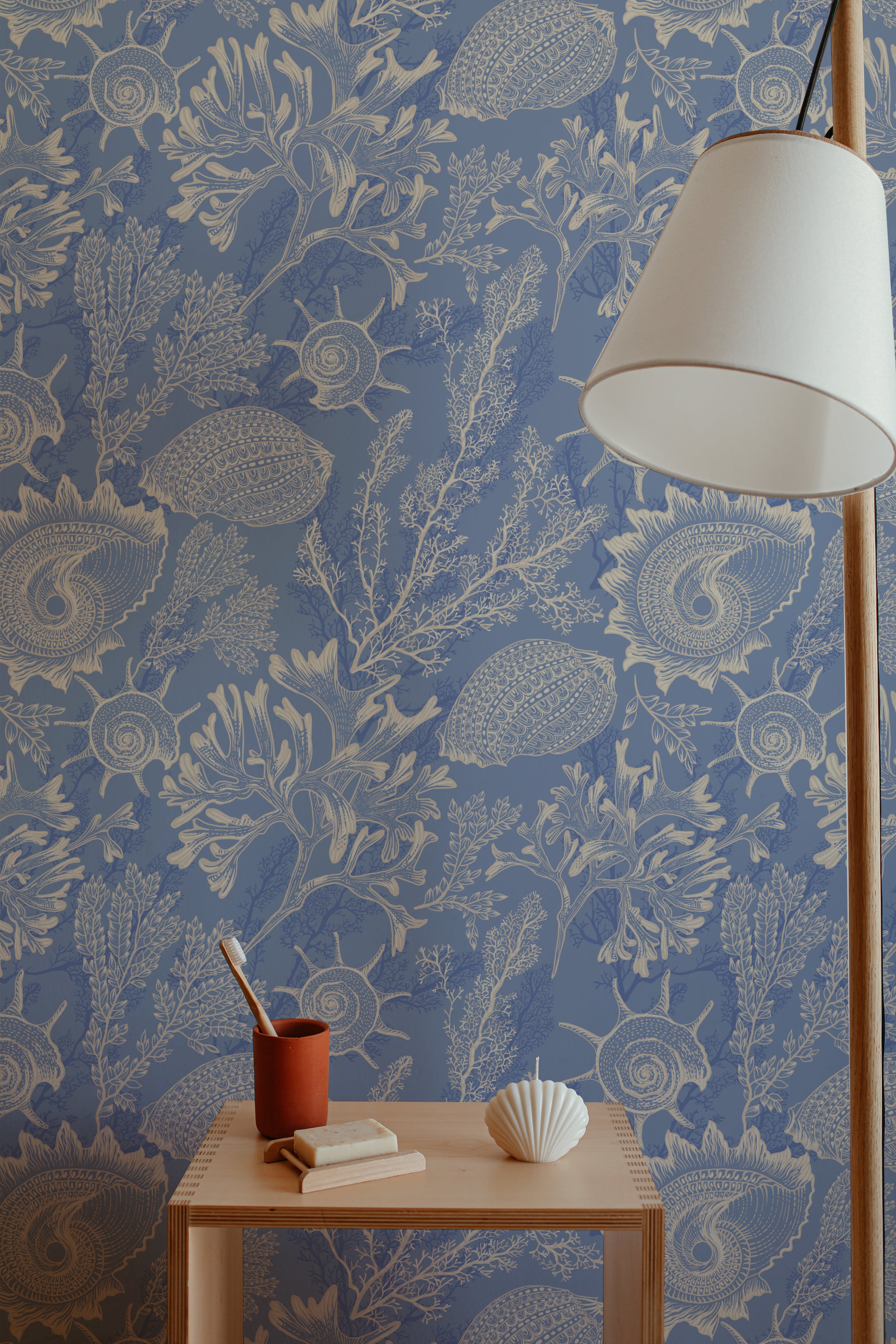 A minimalist interior setup featuring a wooden table with a terracotta cup holding a toothbrush, a bar of soap, and a seashell-shaped candle. A floor lamp with a white shade stands beside the table. The background features Coastal Treasures Wallpaper with intricate illustrations of seashells, coral, and sea plants in beige on a blue background, adding a coastal vibe to the space.