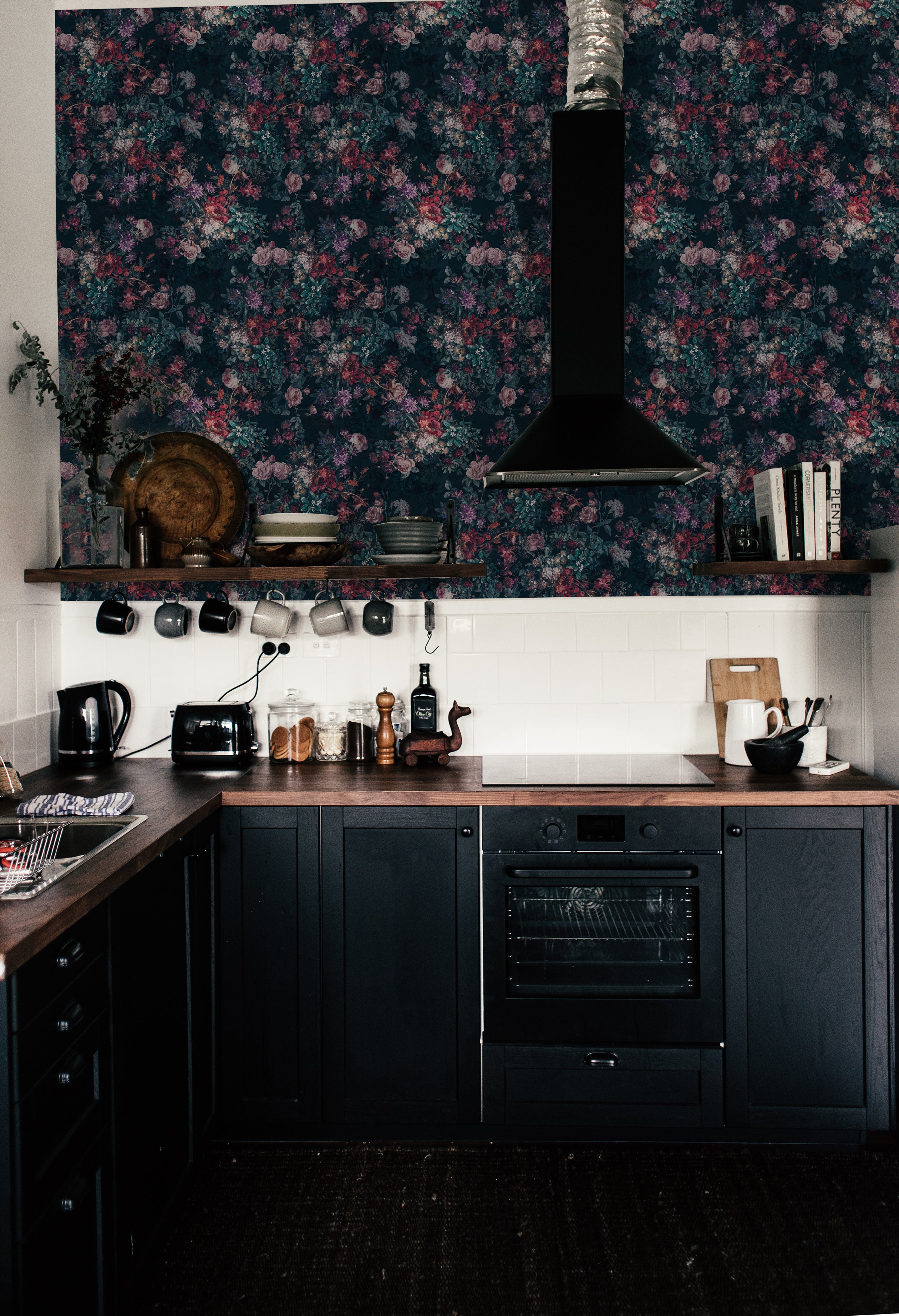 A stylish kitchen featuring the "Dark Luxury Floral Wallpaper - 25" as a backdrop above a white subway tile backsplash. The wallpaper displays a rich, dark floral pattern with dense clusters of roses and other flowers in shades of pink, red, and purple on a deep navy background, complementing the dark wood cabinetry and modern appliances.