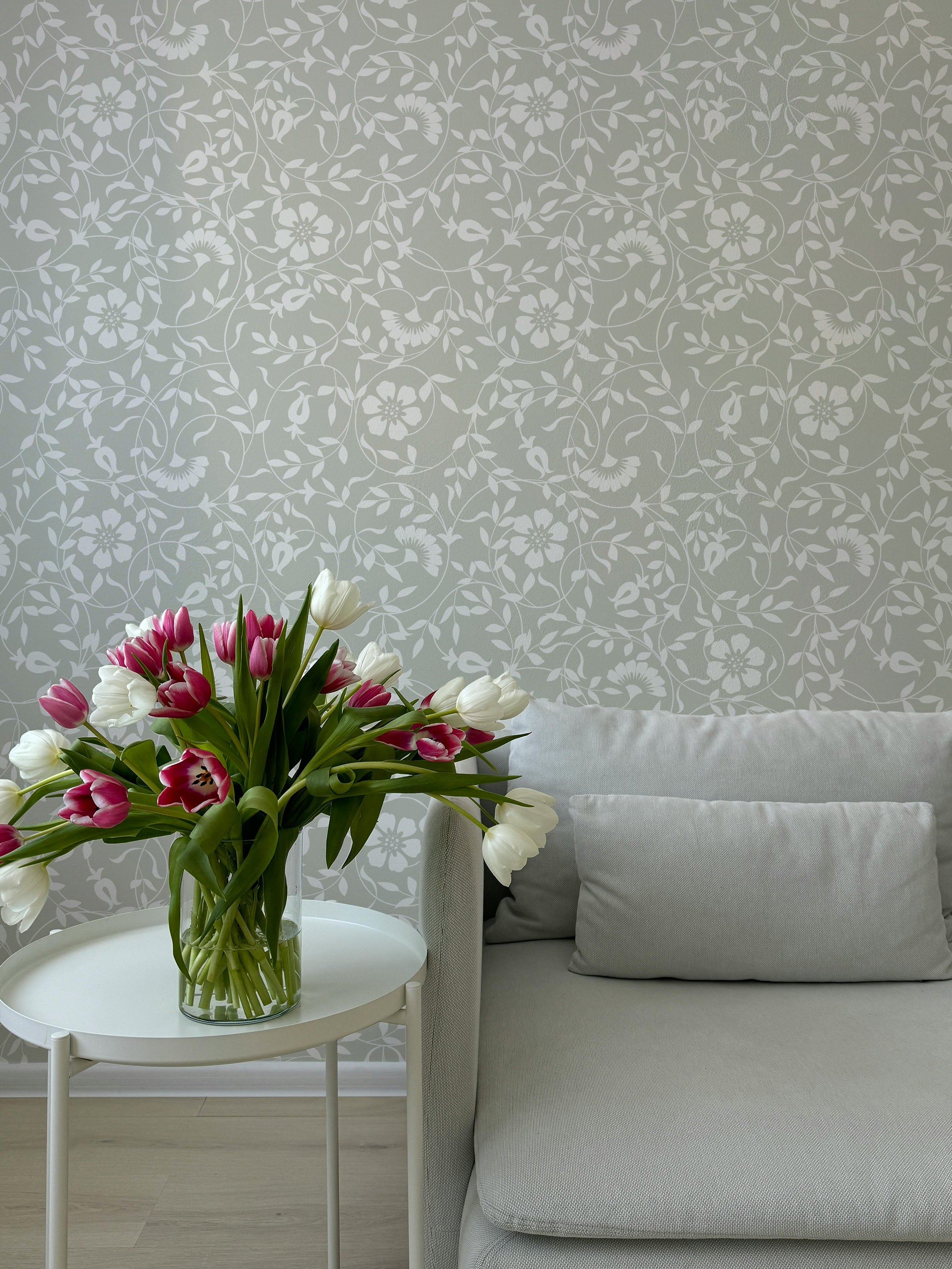 A modern living room setup with a vase of pink and white tulips on a round white table, placed next to a light gray sofa. The background features the Lush Foliage Wallpaper with a delicate floral and leafy pattern in soft green and white, enhancing the room's serene and fresh look.