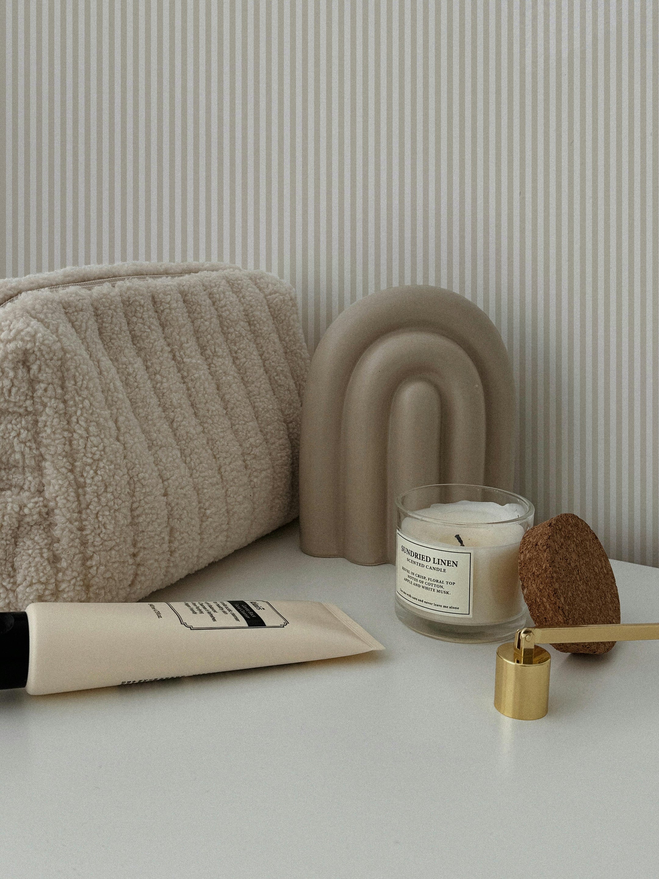 A stylish bathroom counter setup with a textured beige pouch, a minimalist beige arch decor piece, and a candle. The backdrop features the Anne Stripe Wallpaper - Mini with subtle vertical stripes, creating a clean and modern look.