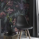 A modern interior with a black chair, a black vase on a gold wire table, and a floor lamp. The wall is adorned with Ephemeral Wallpaper, showcasing a vibrant pattern of large red and pink flowers, green leaves, and purple accents on a dark background.