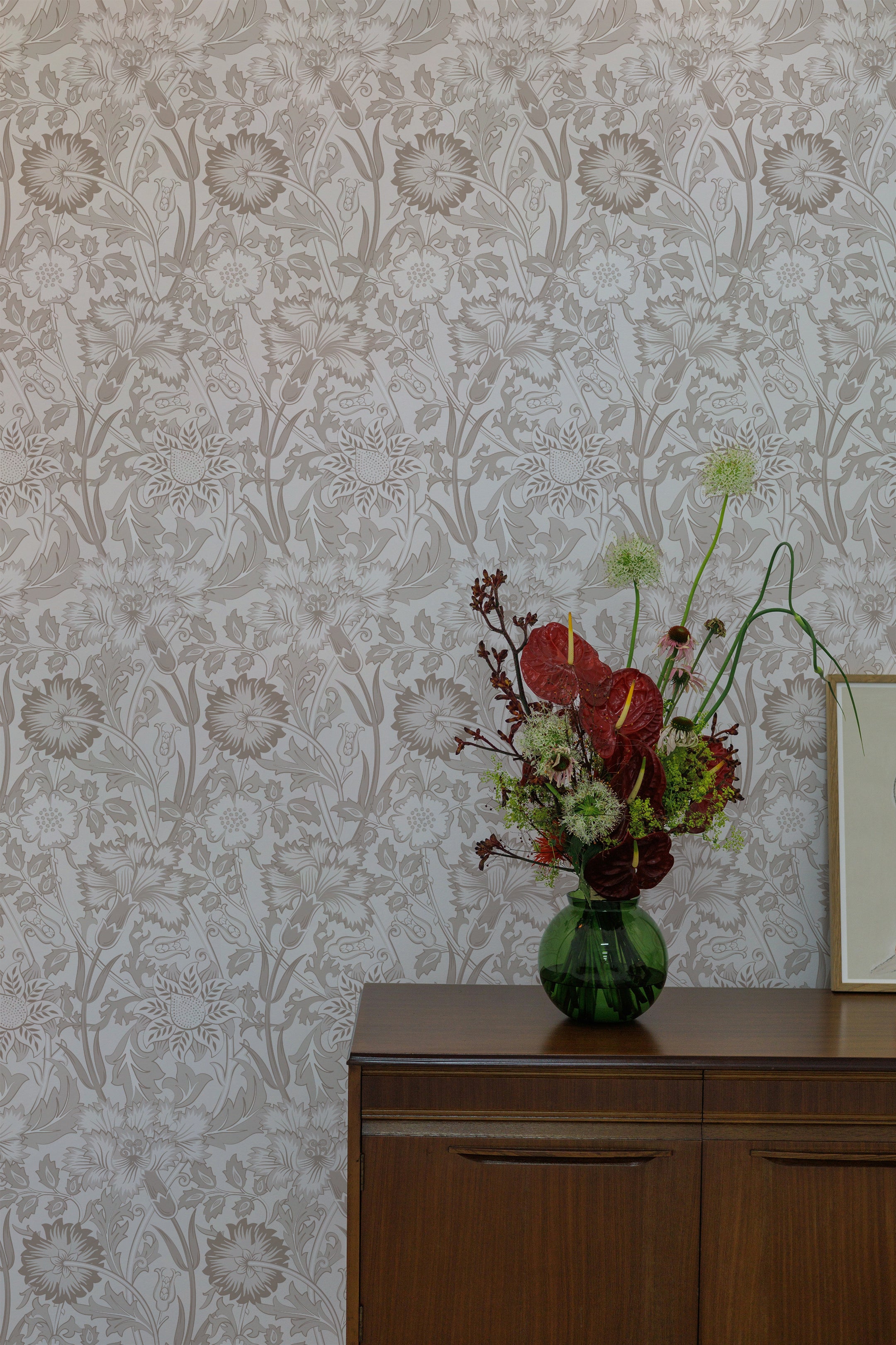 A beautiful floral arrangement in a green glass vase sits on a wooden sideboard, set against a wall covered with Garden Delight wallpaper. The wallpaper's beige floral pattern enhances the natural beauty of the flowers, adding a touch of elegance to the decor.