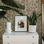 An interior setting showcasing the Little Flowers Wallpaper, with the pattern visible behind a modern white dresser that supports a framed black and white photo, a white vase, and a potted plant.