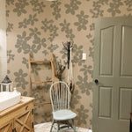 A bathroom beautifully decorated with the Little Flowers Wallpaper. The wallpaper covers one wall, providing a striking contrast to the light fixtures and soft furnishings. This setup includes a vintage wooden vanity, a rustic ladder used as a towel rack, and a simple wooden chair, creating a cozy and inviting space.