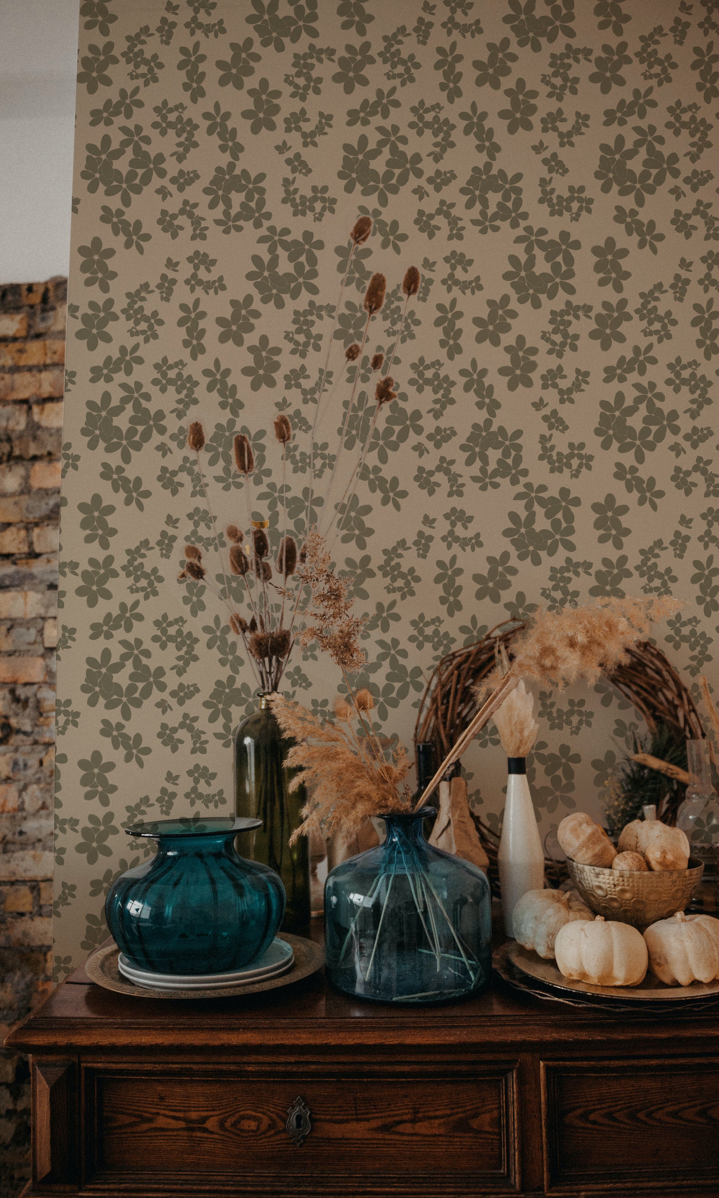 A warm and inviting room decor accentuated with the Little Flowers Wallpaper. The room features a vintage wooden dresser adorned with autumn-themed decorations including dried flowers, pumpkins, and colorful vases. The floral wallpaper adds a soft, botanical backdrop that complements the rustic and seasonal elements of the room's decor.