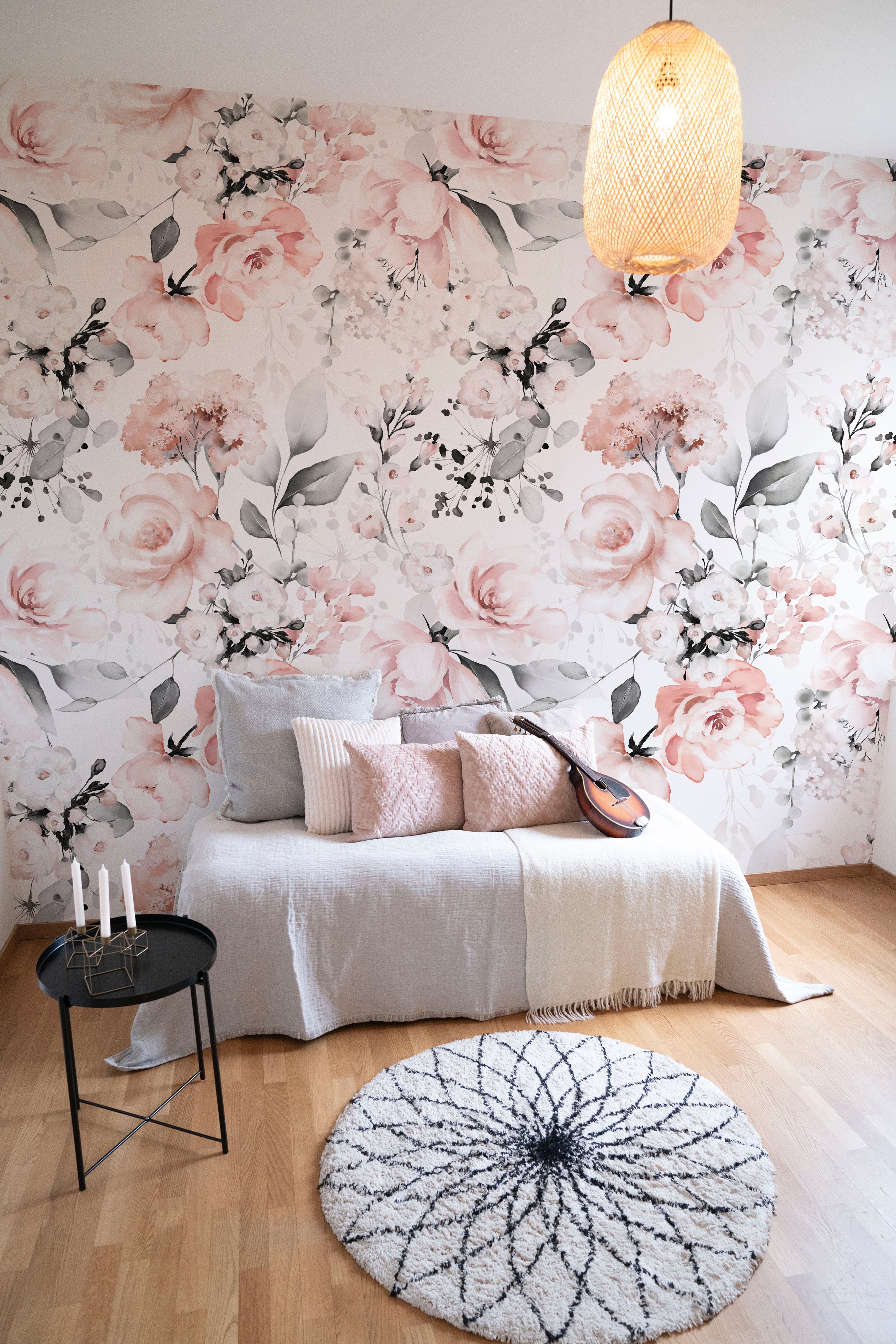 A cozy living room with a floral wallpaper accent wall. The wallpaper features a delicate pattern of pink flowers and grey leaves on a white background. The room includes a comfortable sofa with pastel cushions, a round rug, a small black side table with candles, and a hanging wicker lamp
