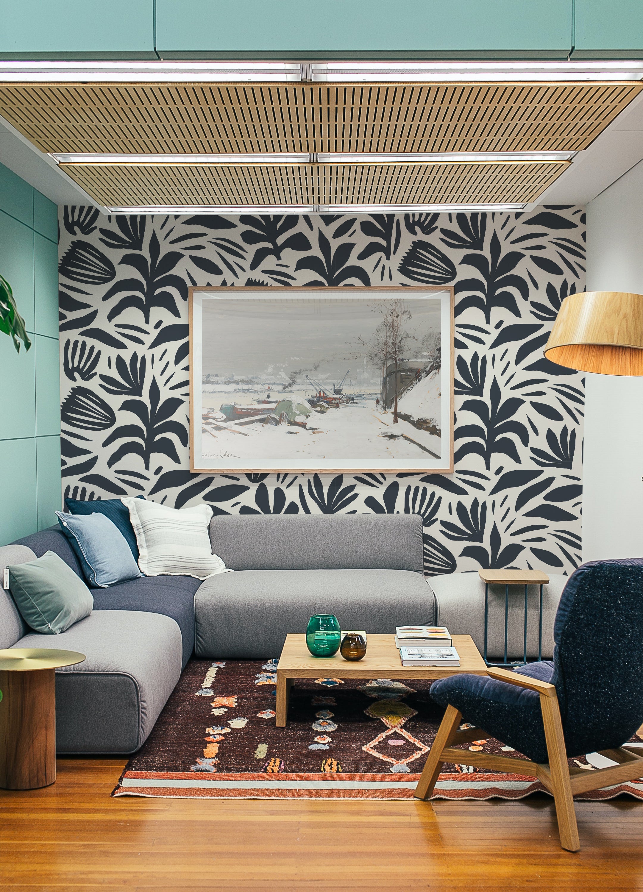 A modern living room with a grey sectional sofa, a colorful patterned rug, and a wooden coffee table. The wall behind the sofa is adorned with Dark Bloom Wallpaper, showcasing bold black abstract floral and leaf patterns on a white background, complemented by a framed landscape artwork.