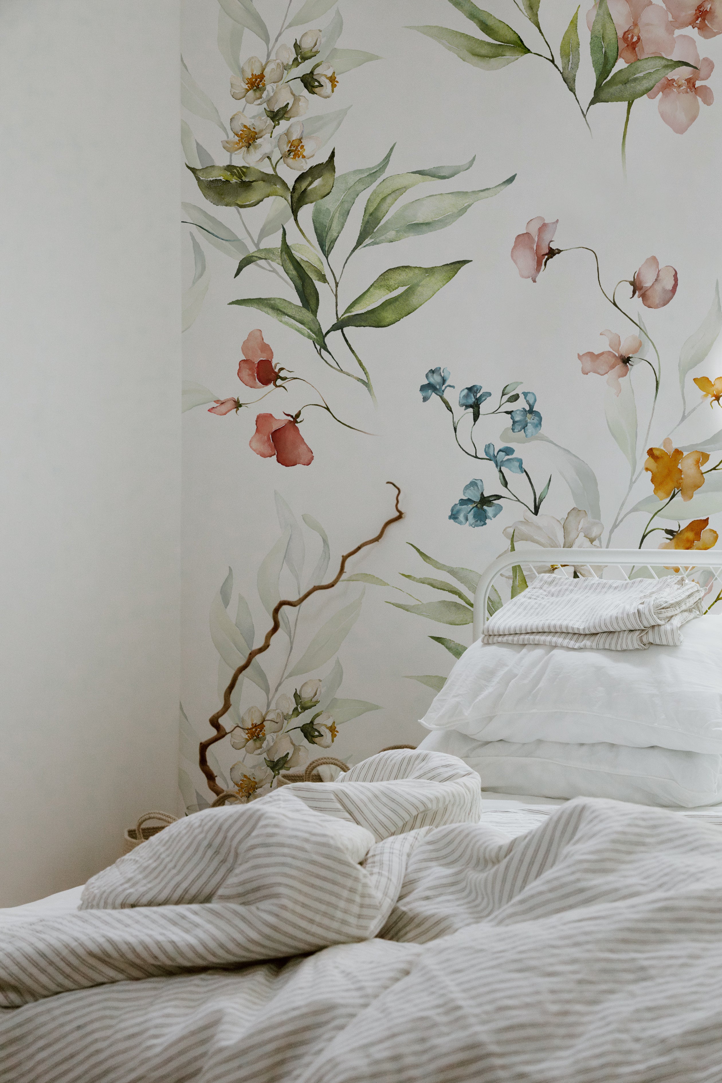 A bedroom wall adorned with a floral wallpaper design showcasing watercolor illustrations of flowers and leaves. The pattern features soft pink, blue, yellow, and white flowers with green foliage, adding a touch of nature to the room's decor. The bed in the foreground has white linens and striped bedding