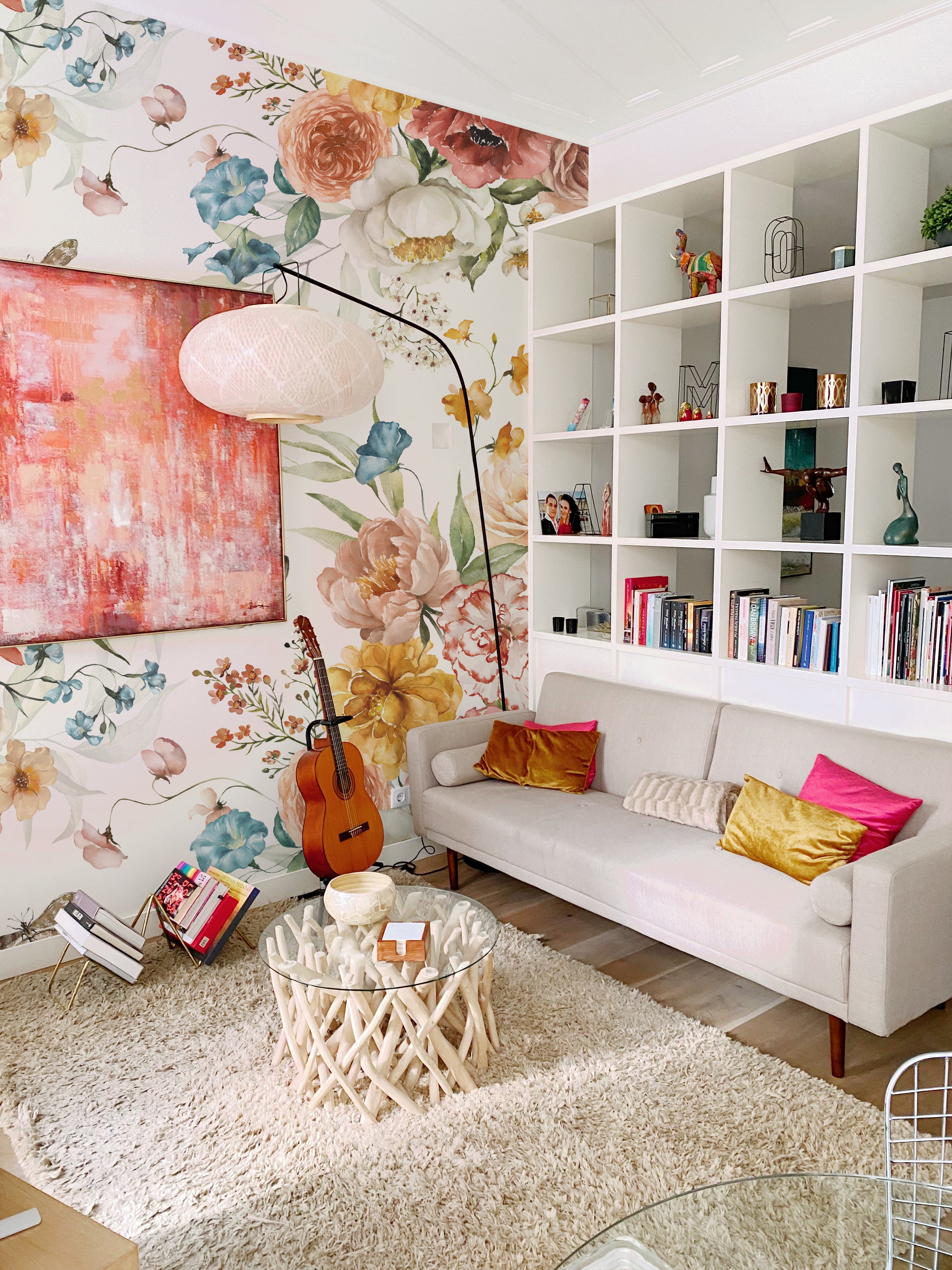 A cozy living room with a floral wallpaper that features a vibrant watercolor design of roses, peonies, and other flowers in red, pink, yellow, blue, and white, along with green foliage. The room includes a white sofa with colorful cushions, a guitar, a glass coffee table with a natural wood base, and a bookshelf filled with books and decor items