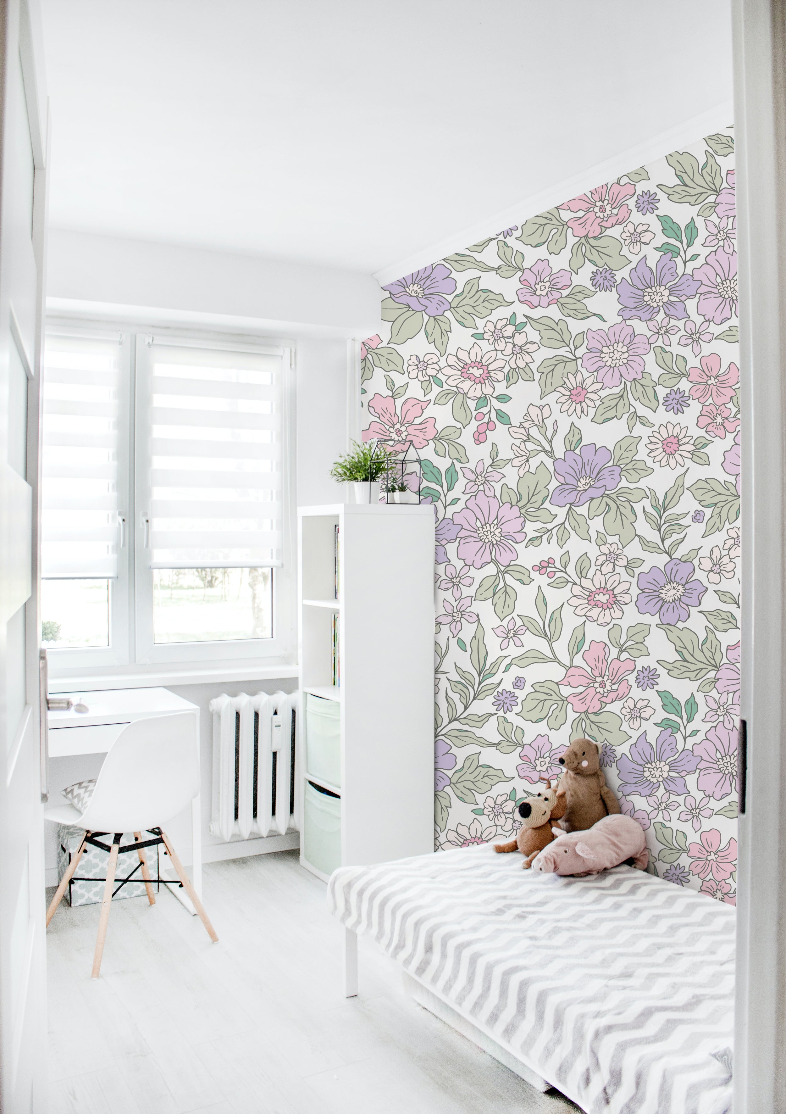 A bright and airy bedroom with a floral wallpaper accent wall. The wallpaper features a whimsical pattern of pastel-colored flowers in pink, lavender, and green. The room includes a small white desk, a bookshelf, and a bed with striped bedding, adorned with plush toys.