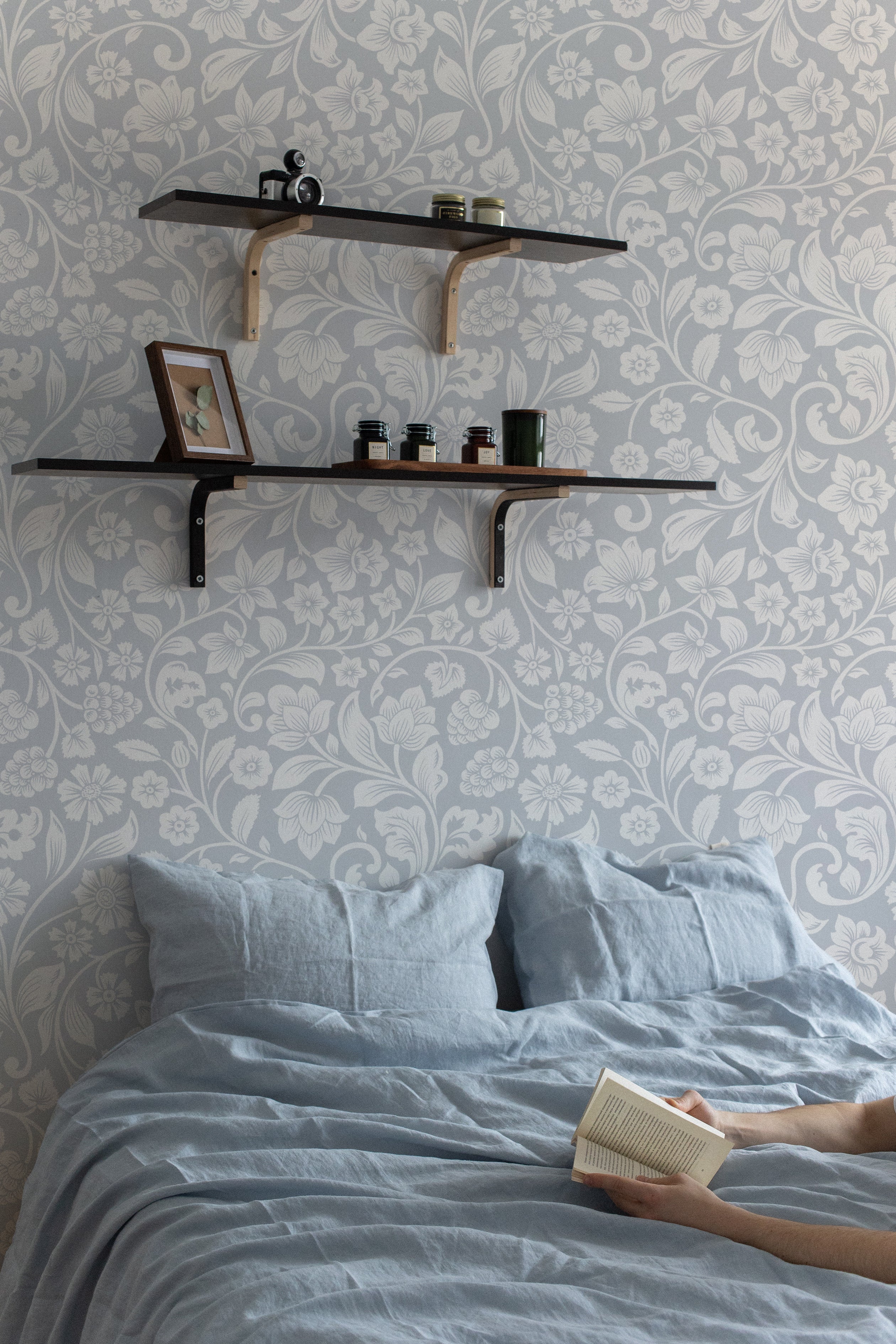 Faded Blue Vintage Floral Wallpaper - 75" used in a cozy bedroom setting with floating shelves and blue bed linens, highlighting the elegant floral pattern.