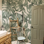 A stylish bathroom adorned with the Modern Monstera Wallpaper creating a bold and tropical ambiance. The large monstera leaf pattern dominates the space, complementing the wooden bathroom vanity and a light blue chair. Accessories like a woven basket and rustic elements enhance the natural, organic feel of the room.