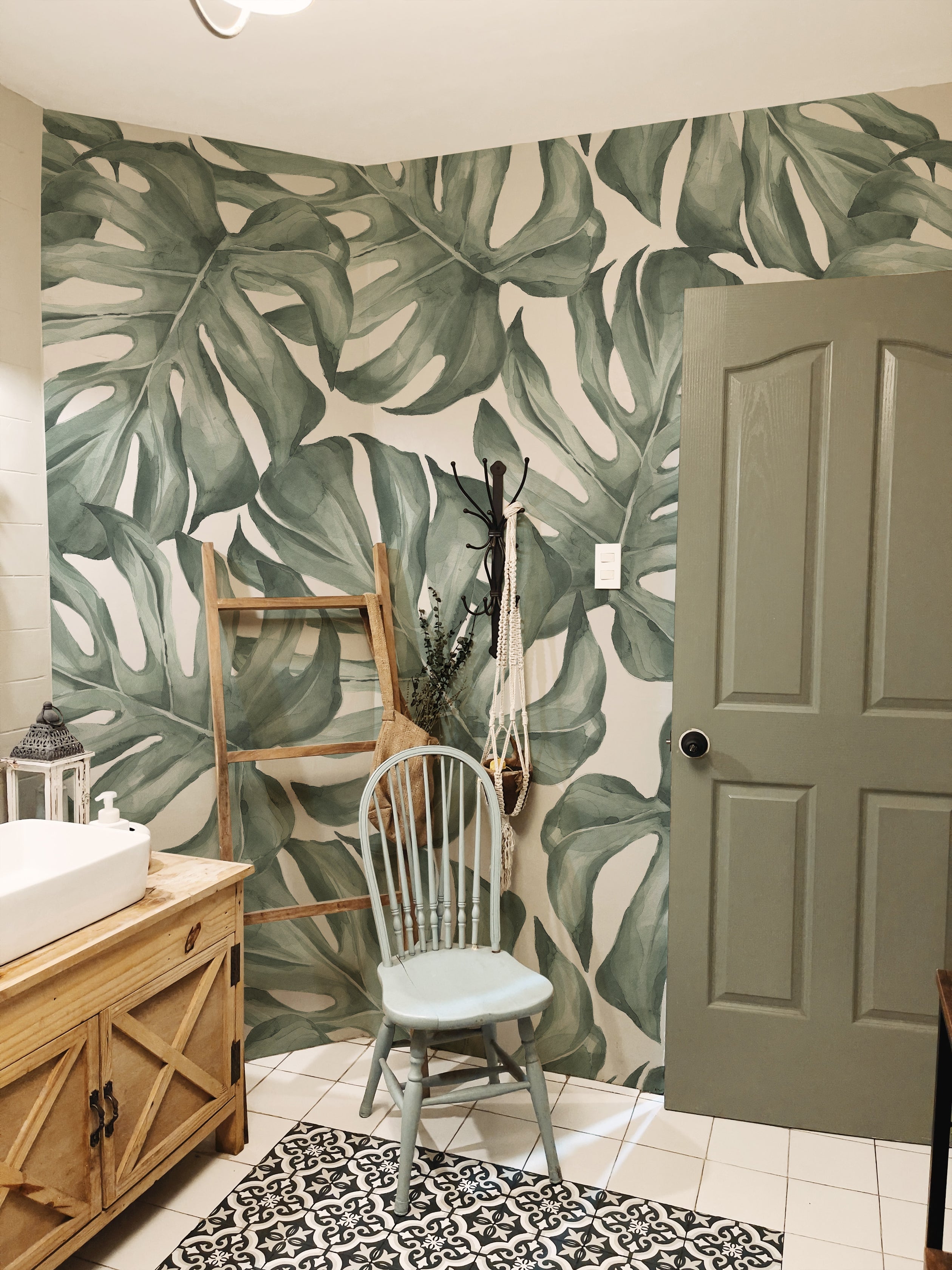 A stylish bathroom adorned with the Modern Monstera Wallpaper creating a bold and tropical ambiance. The large monstera leaf pattern dominates the space, complementing the wooden bathroom vanity and a light blue chair. Accessories like a woven basket and rustic elements enhance the natural, organic feel of the room.