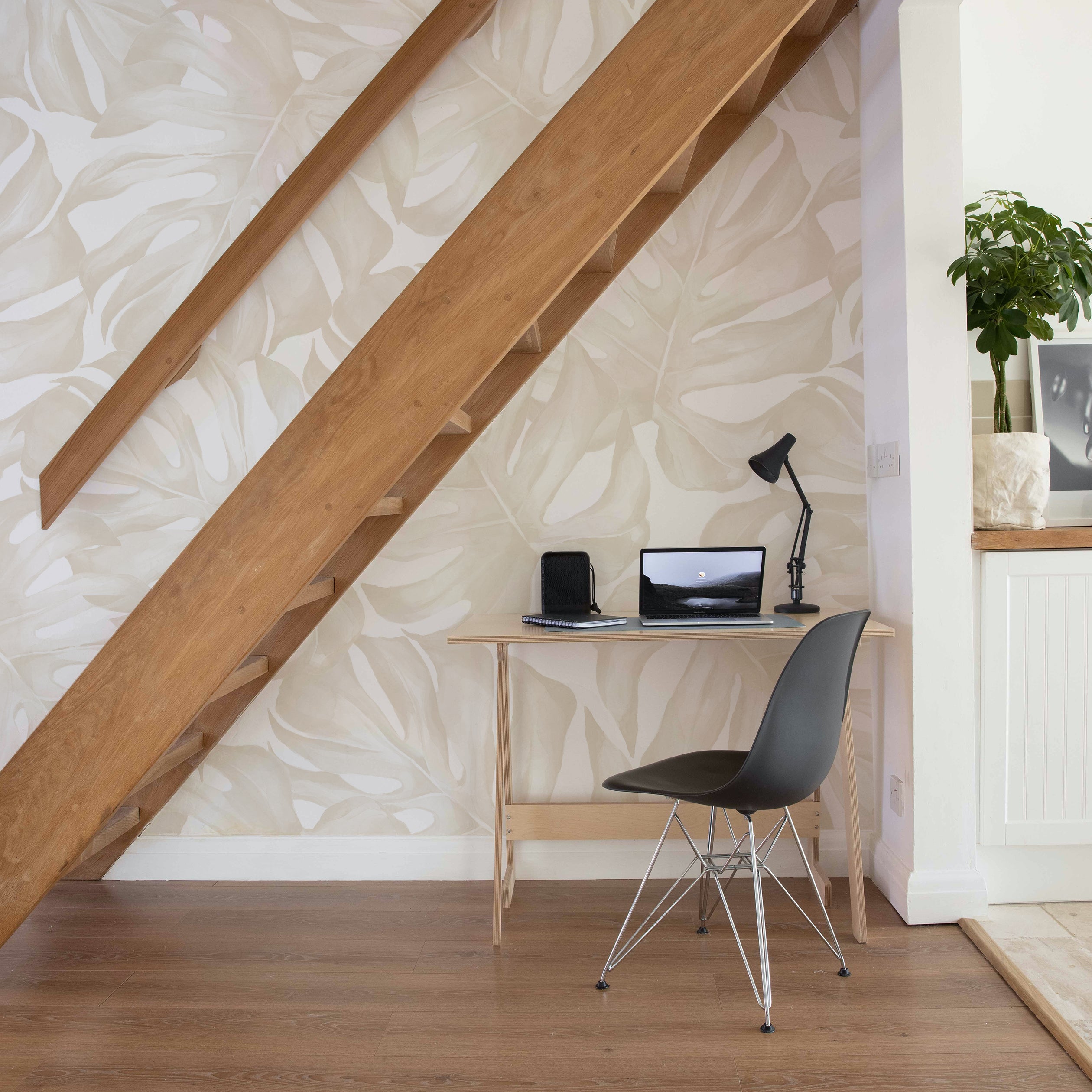 Interior view of a home office setup with Modern Oasis Wallpaper - Ecru. The wallpaper features a subtle, large-scale monstera leaf pattern in ecru tones, providing a calming and sophisticated atmosphere under a wooden staircase.