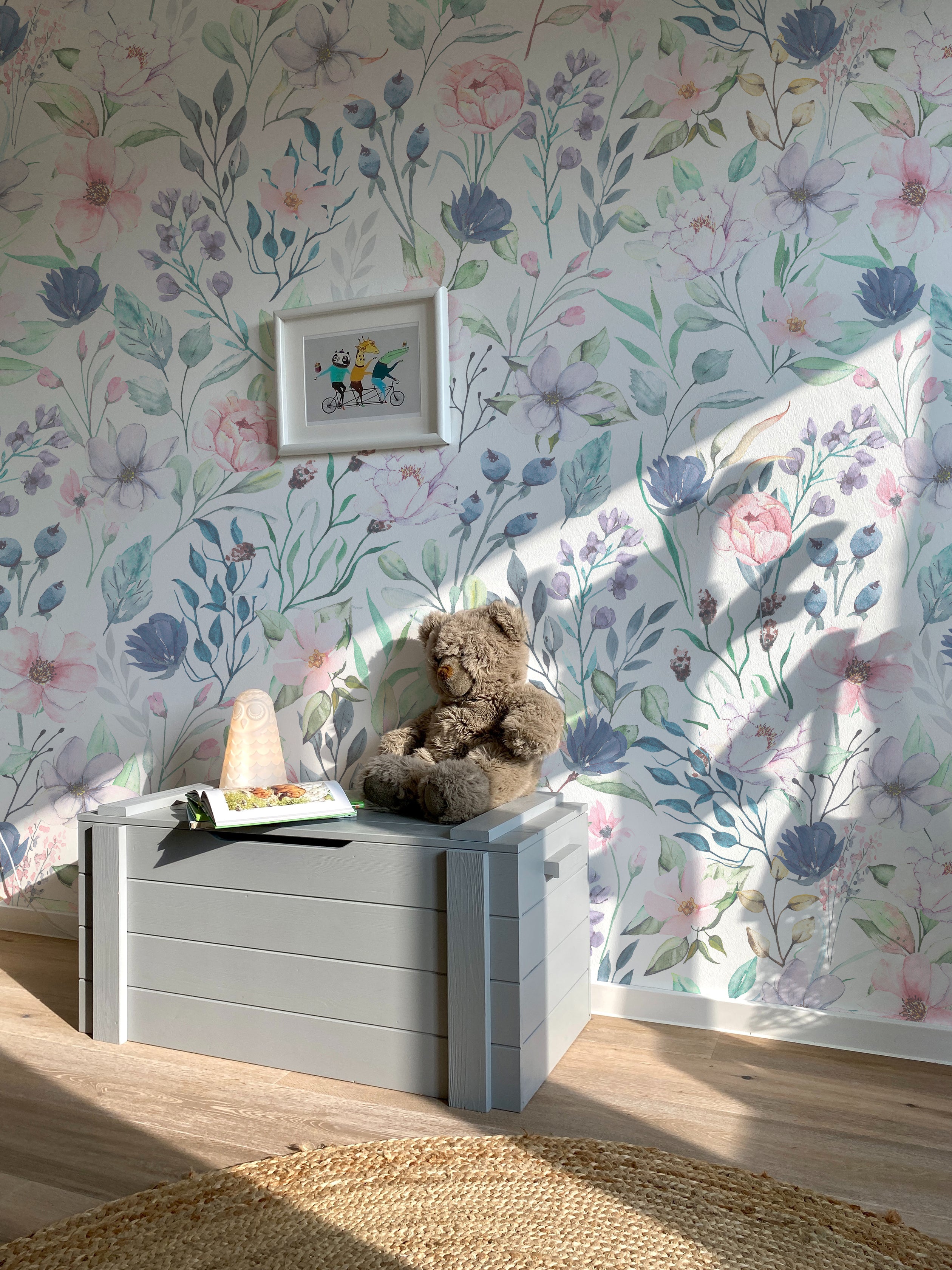 The wallpaper is installed in a child's room, creating a serene and inviting atmosphere with its delicate floral design. A teddy bear sits on a grey toy chest in front of the wallpaper.
