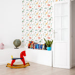 A child's playroom featuring a wall covered in Dinosaur Nursery Wallpaper. The design includes playful dinosaurs, tropical plants, and abstract elements in pastel shades. Decorative elements such as a red rocking horse, a globe, and a bookshelf enhance the educational and playful environment.
