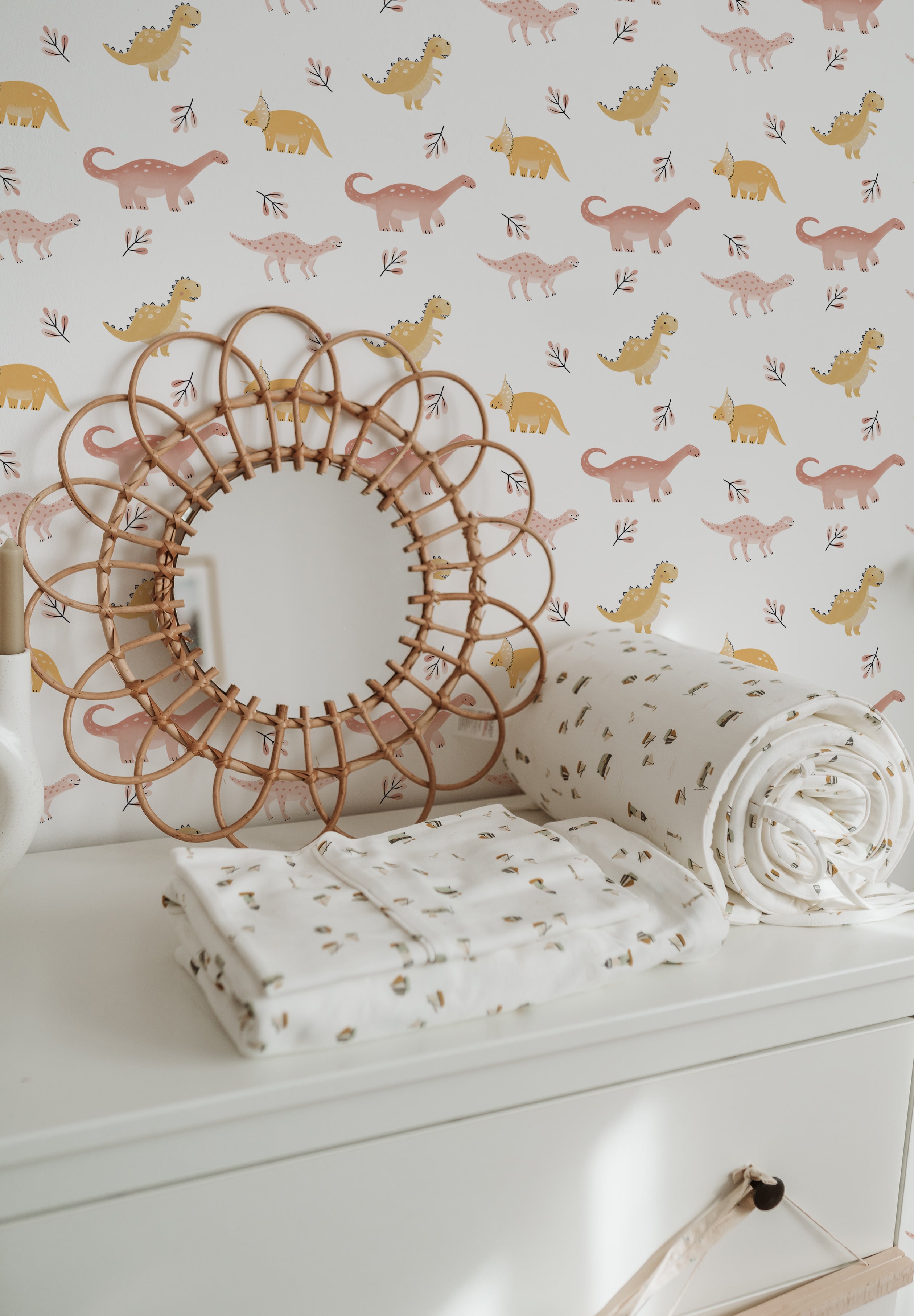 A child's room setup showcasing the "Dino Days Wallpaper III" on one wall, with coordinating decor. The room includes a white dresser topped with a decorative rattan mirror and rolled-up dinosaur-themed wallpapers, complementing the playful and colorful dinosaur patterns on the wall.