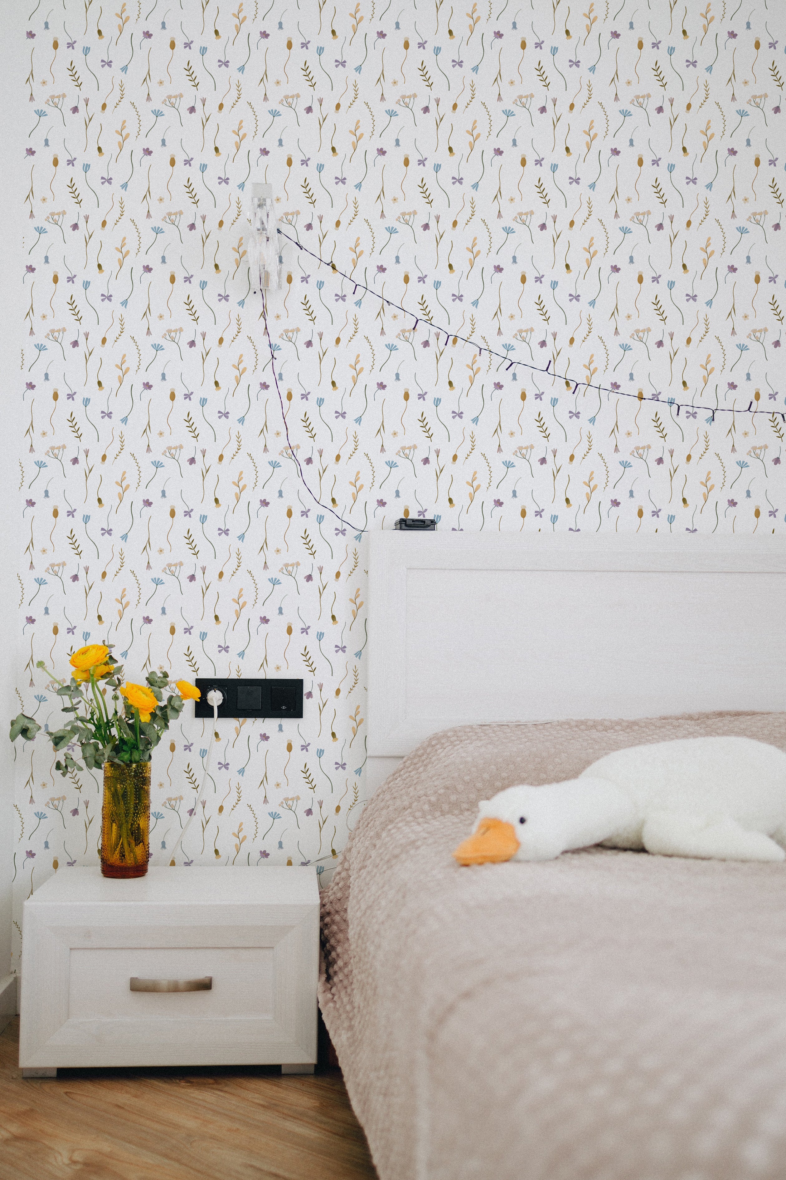 A cozy bedroom setting featuring the Minimal Paradise Wallpaper. The wallpaper adds a subtle and elegant backdrop to the room, complemented by simple decor including a wooden bedside table with a lamp, a vase of fresh flowers, and a white plush toy. The wallpaper’s understated botanical design enhances the room's calm and peaceful ambiance.