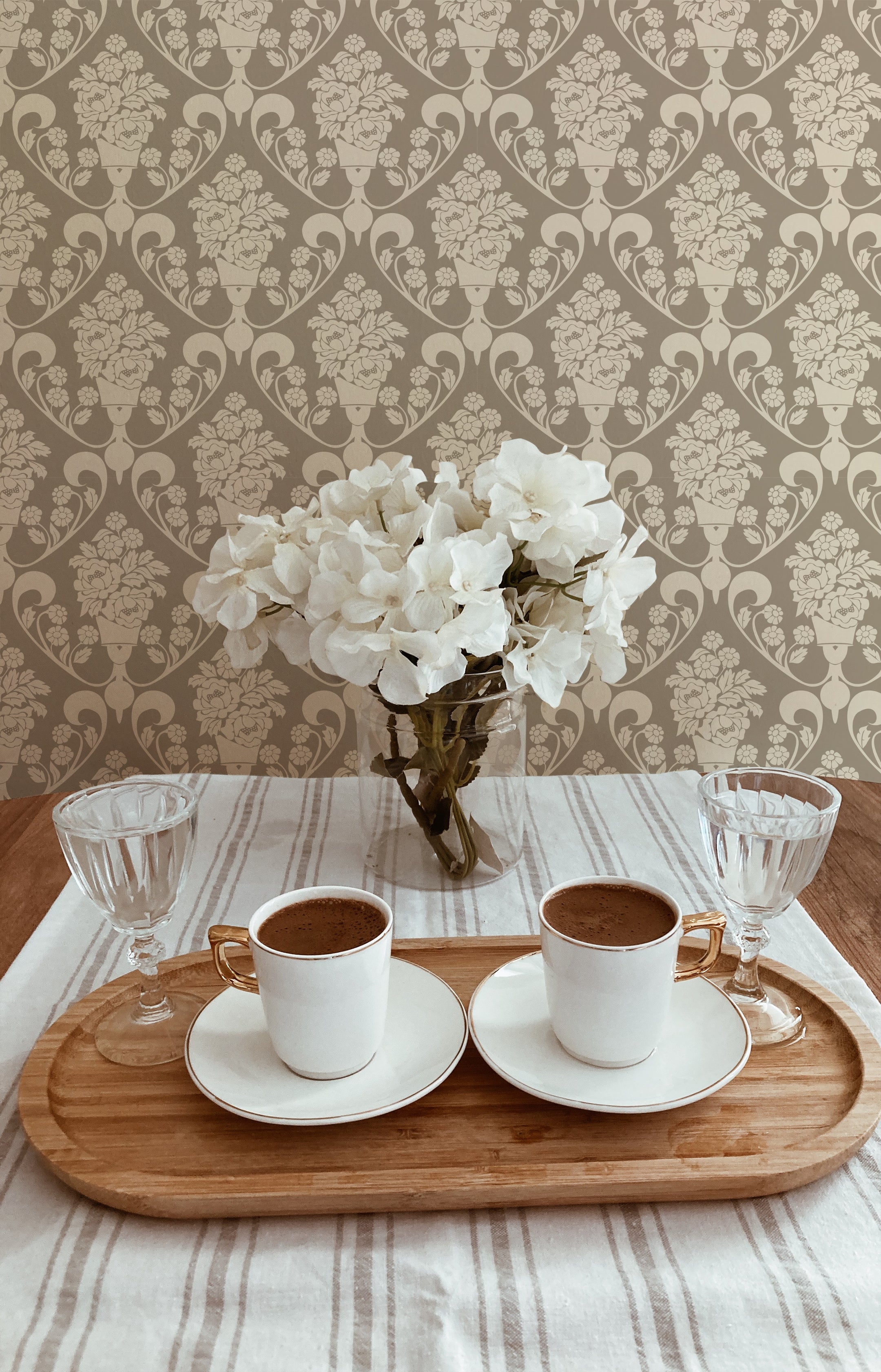 A quaint coffee table setting with two white coffee cups on a wooden tray, placed in front of a wall covered with Victorian Bloom wallpaper. The wallpaper's beige floral pattern complements the fresh white flowers in a glass vase, creating a serene and inviting atmosphere.