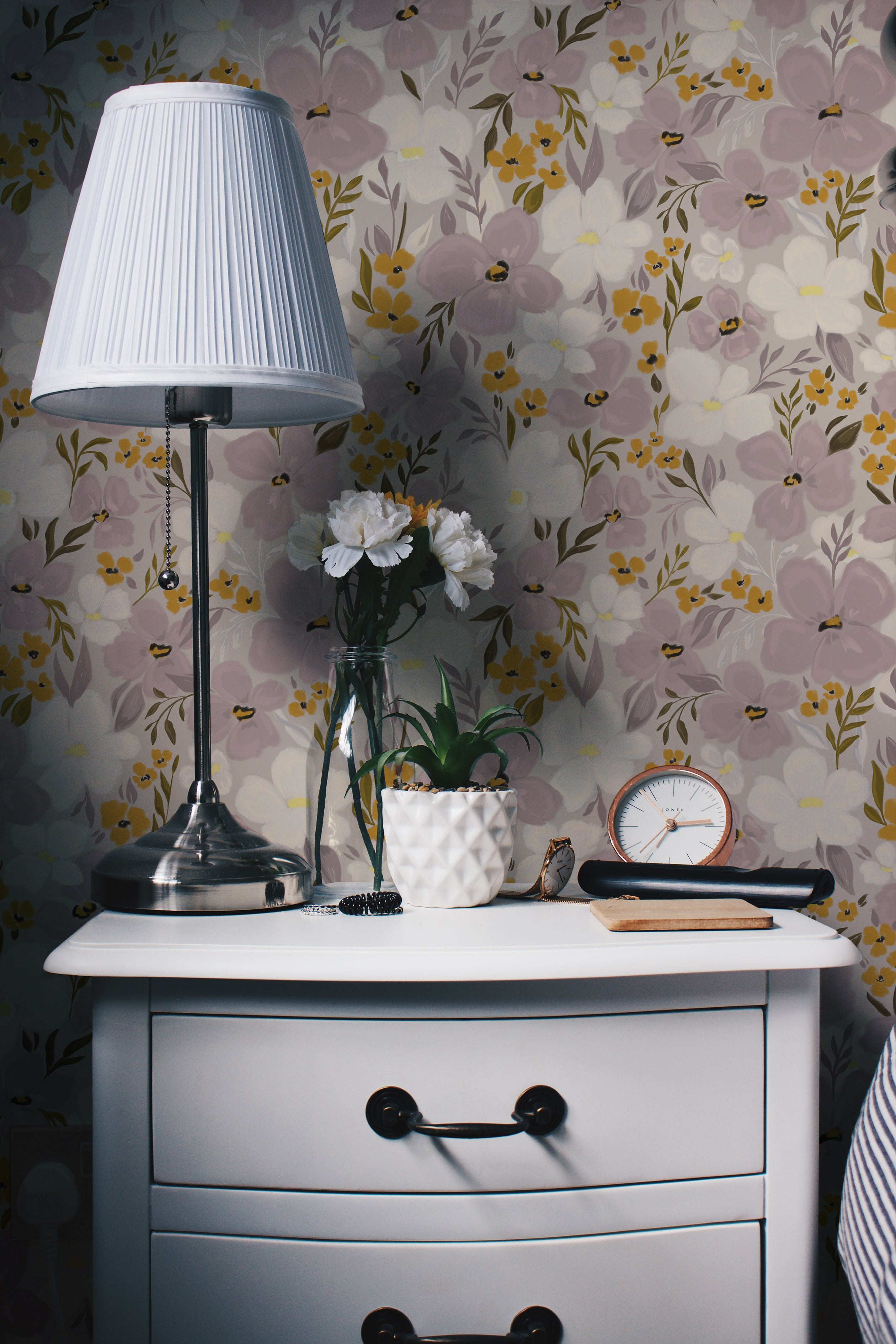 An interior decor setting displaying the Fleur Wallpaper as a background, featuring a white dresser with a silver lamp, white flower vase, and various small items, enhancing the room's aesthetic
