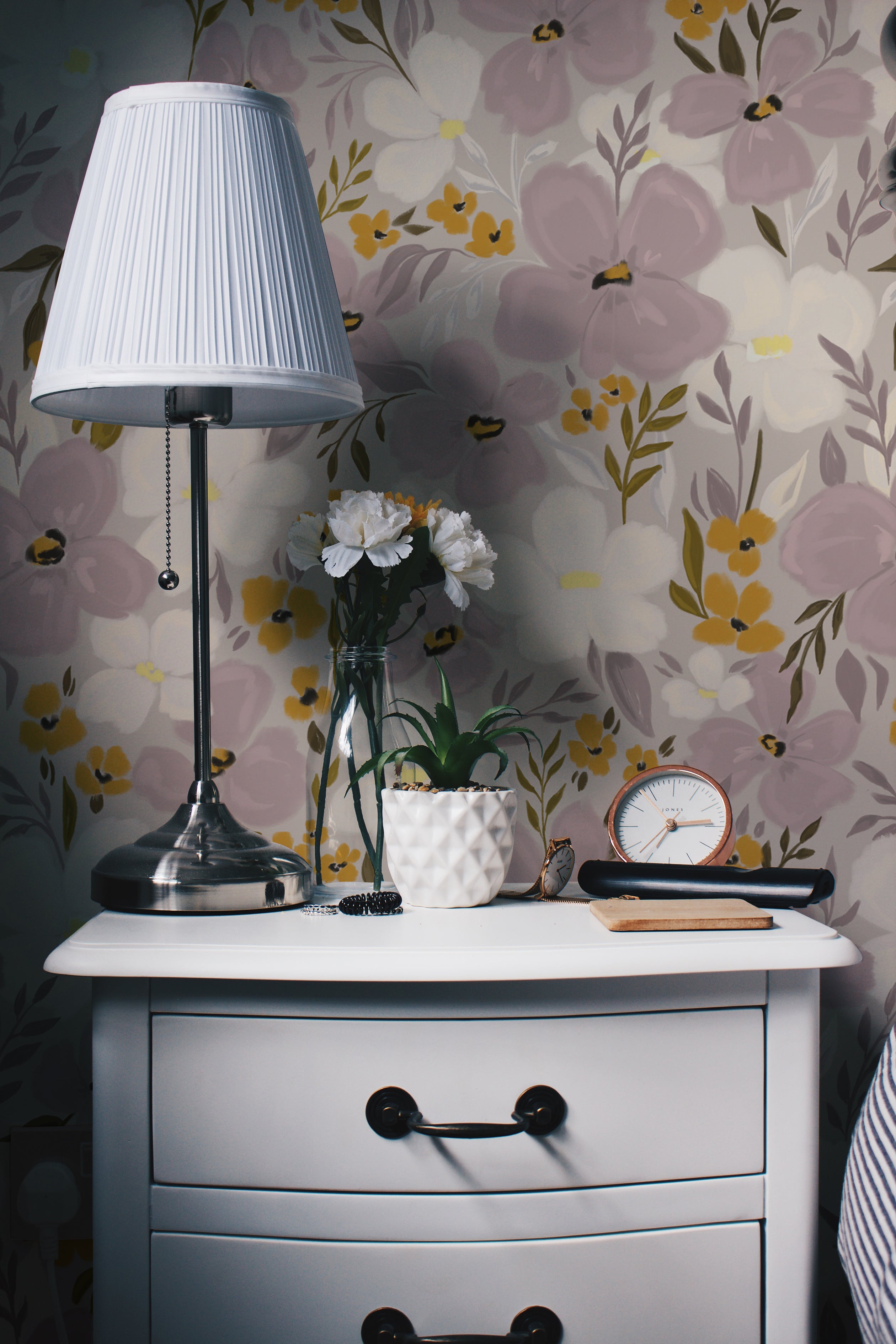 An elegantly decorated space showcasing the Fleur Wallpaper. The wallpaper provides a charming backdrop to a white nightstand, topped with a silver lamp, a glass vase with white flowers, a succulent plant in a ceramic pot, and a vintage clock. The floral pattern adds a fresh and airy feel to the room, enhancing the light and cozy decor.