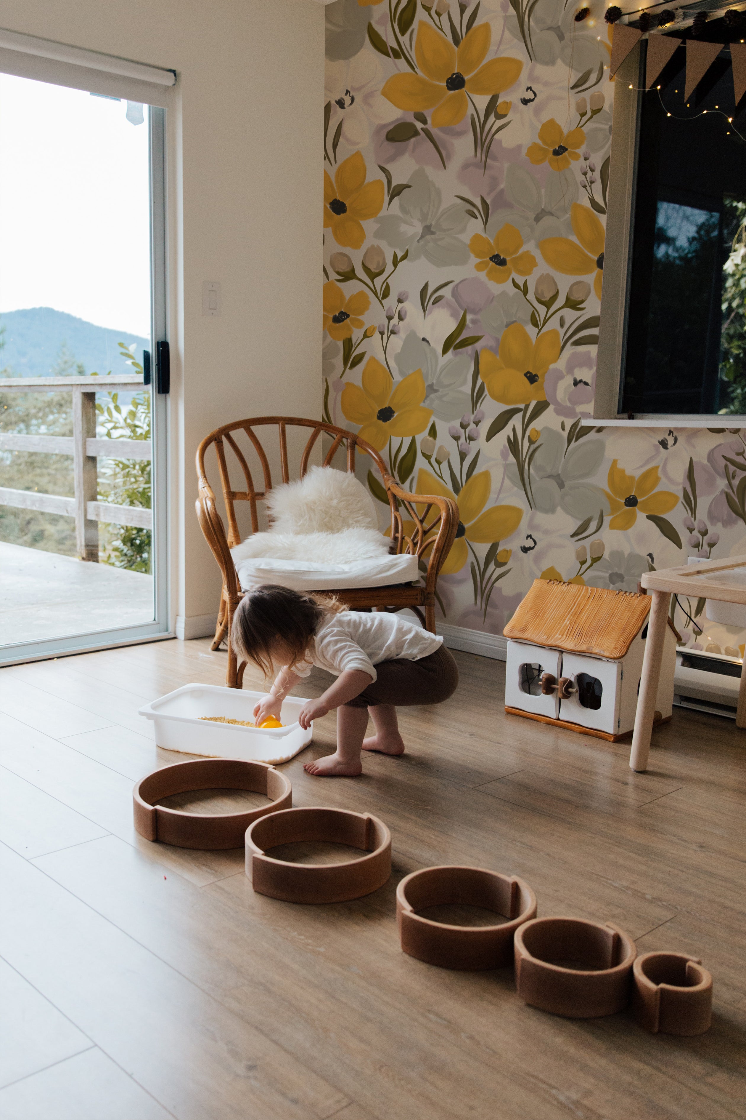 A child's play area with the Yellow Fleur Wallpaper - 75" adorning the wall, adding a cheerful burst of yellow florals to the space, complemented by natural light, wooden toys, and a rattan chair with a white cushion, creating an inviting and playful environment.