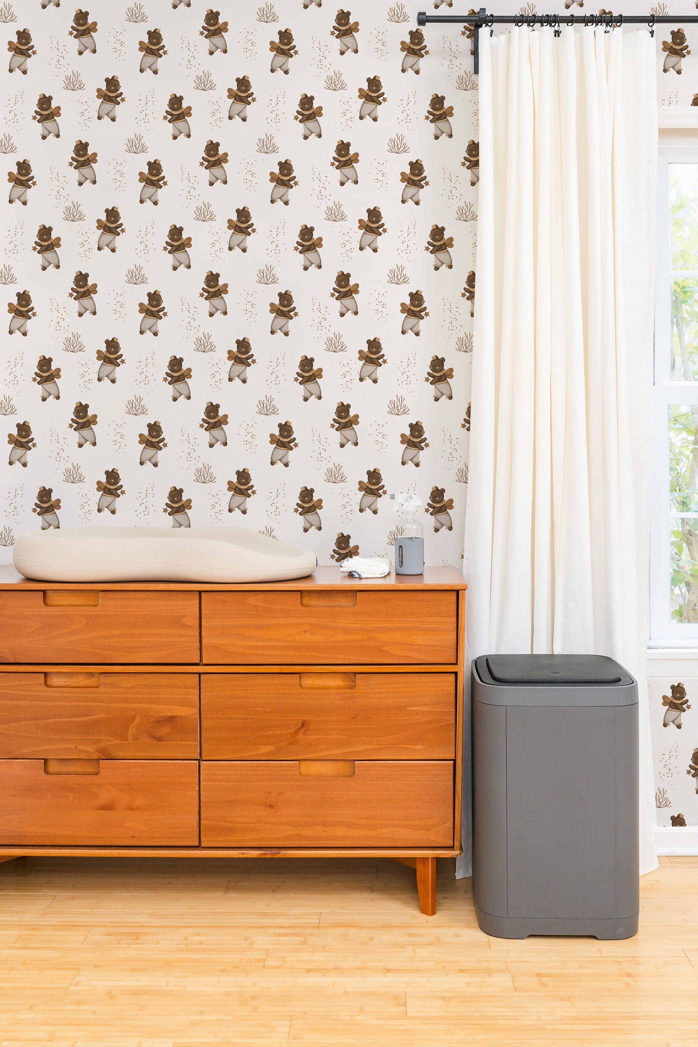 A cozy nursery featuring Stardust Bears Wallpaper with whimsical bears wearing crowns and wings. A wooden dresser and soft lighting create a warm atmosphere.