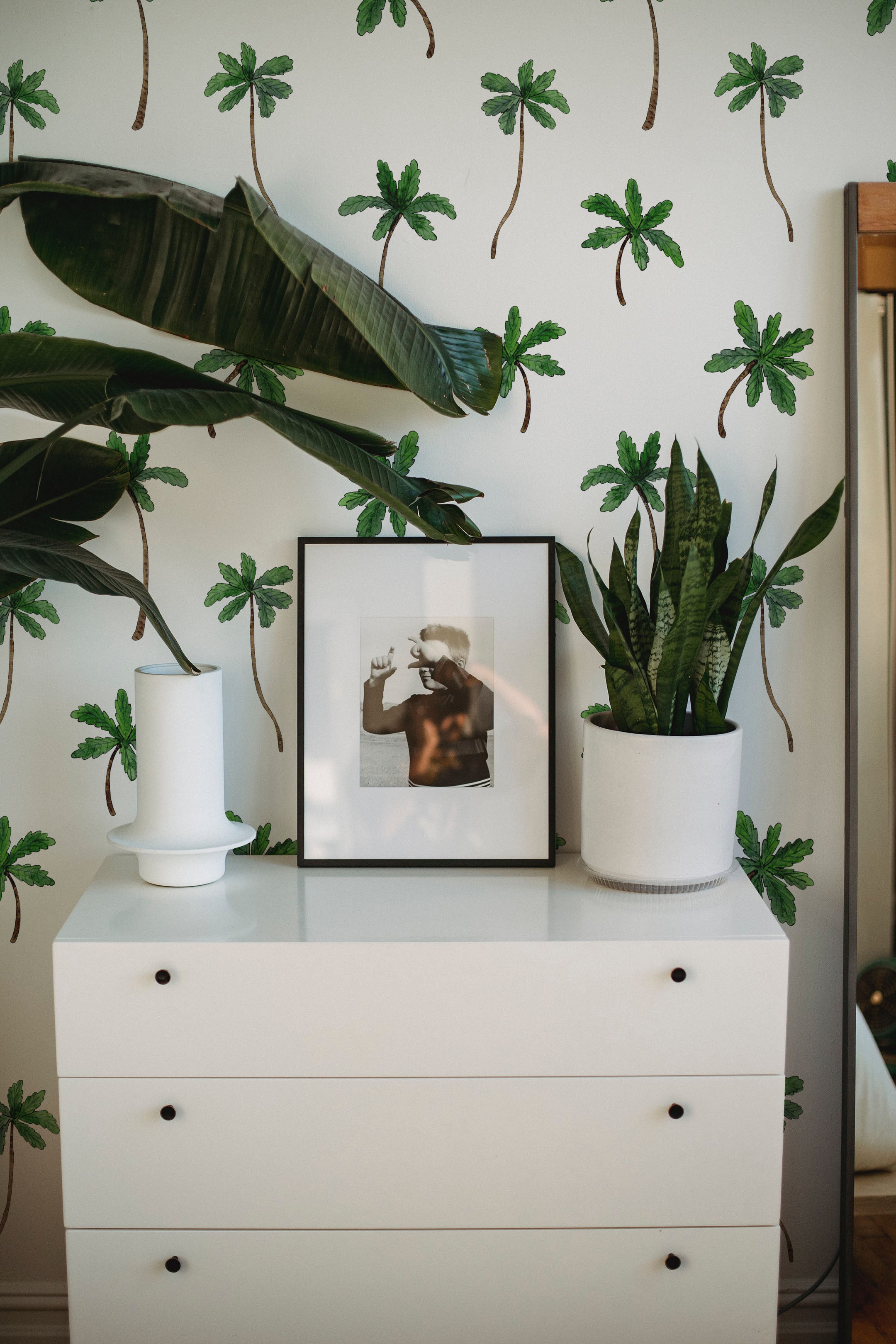 A stylish dresser area with Island Breeze Wallpaper in the background, adorned with green palm trees. The space features a white dresser, framed artwork, and lush plants.
