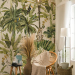 A cozy corner room showcasing the same tropical wallpaper with palm trees and animals. The room is styled with natural decor including a rattan chair, a round woven rug, tall baskets, a side table with books and a vintage camera. Pendant lights hang from the ceiling, casting a warm glow that complements the naturalistic theme.