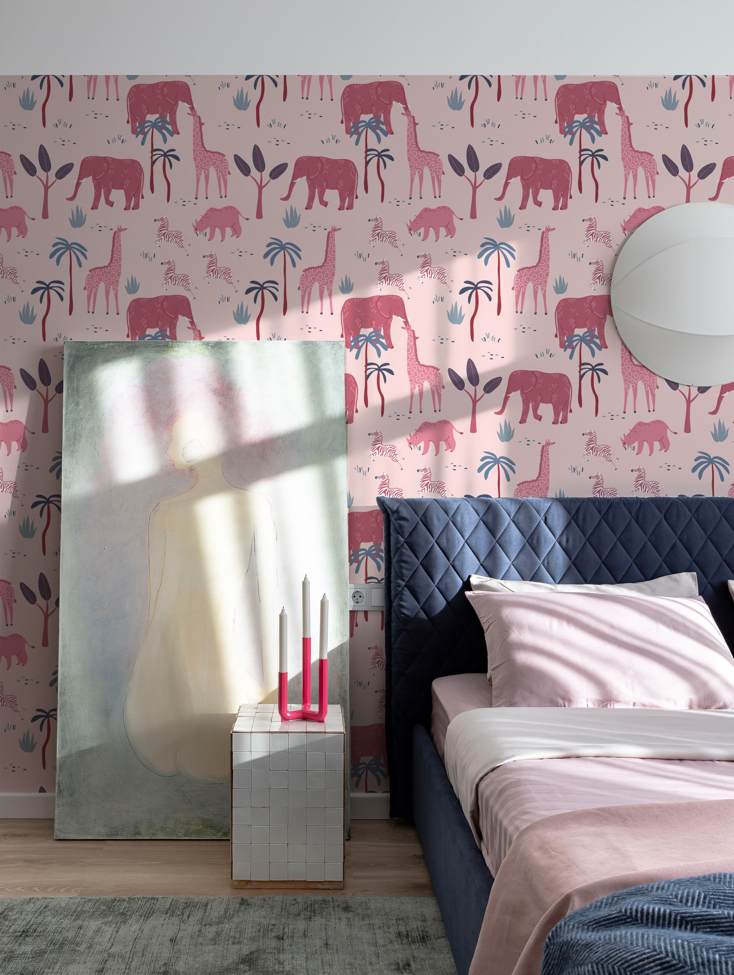 A modern bedroom featuring pink safari-themed wallpaper with elephants, giraffes, and zebras. The room has a dark blue quilted headboard and a minimalist nightstand with pink and white decor.