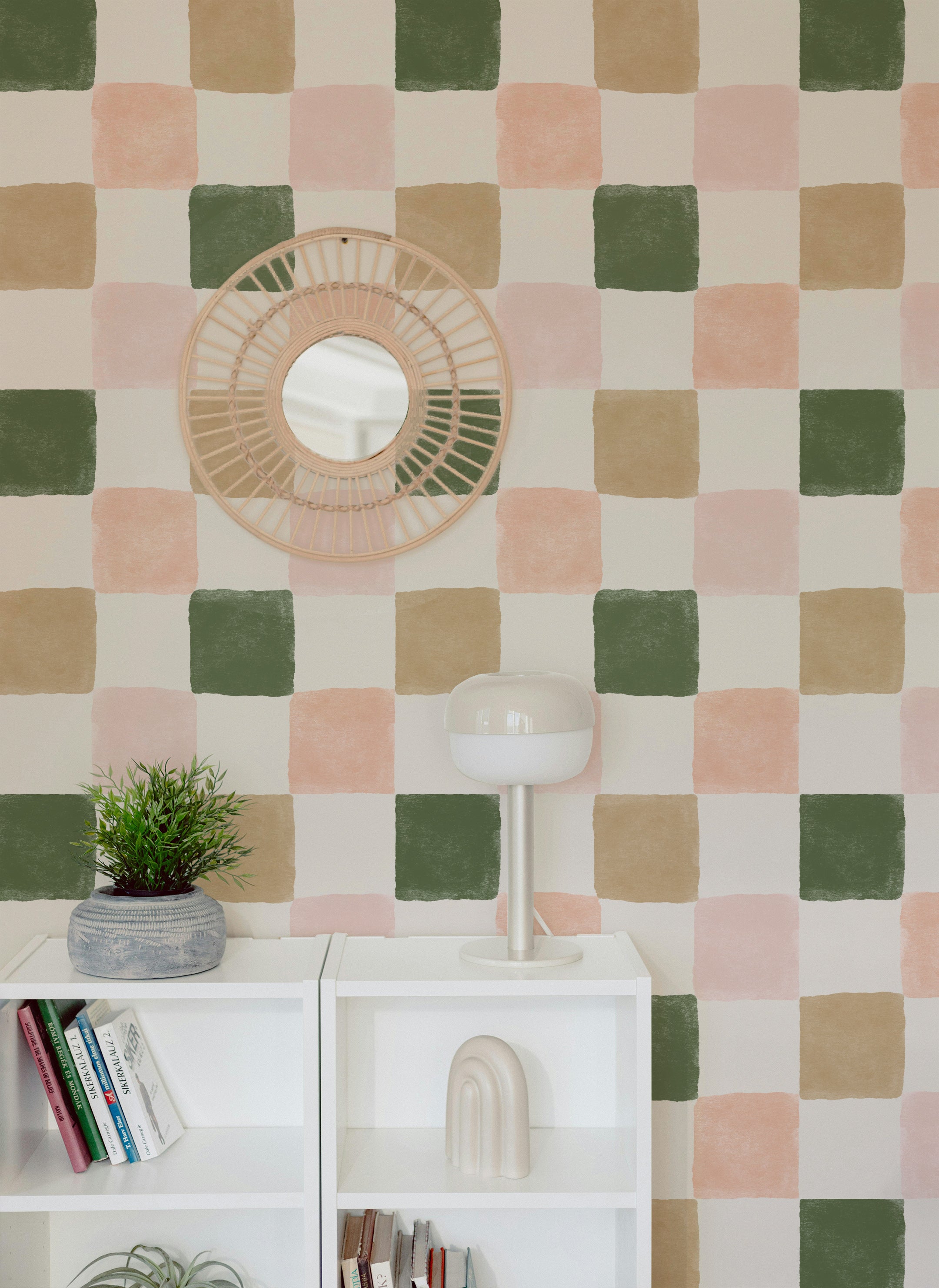 A stylish interior decorated with Clémence Wallpaper II, featuring a checkered pattern in pastel shades of pink, green, and beige. The room includes white shelves, a potted plant, a modern lamp, and a round woven mirror.