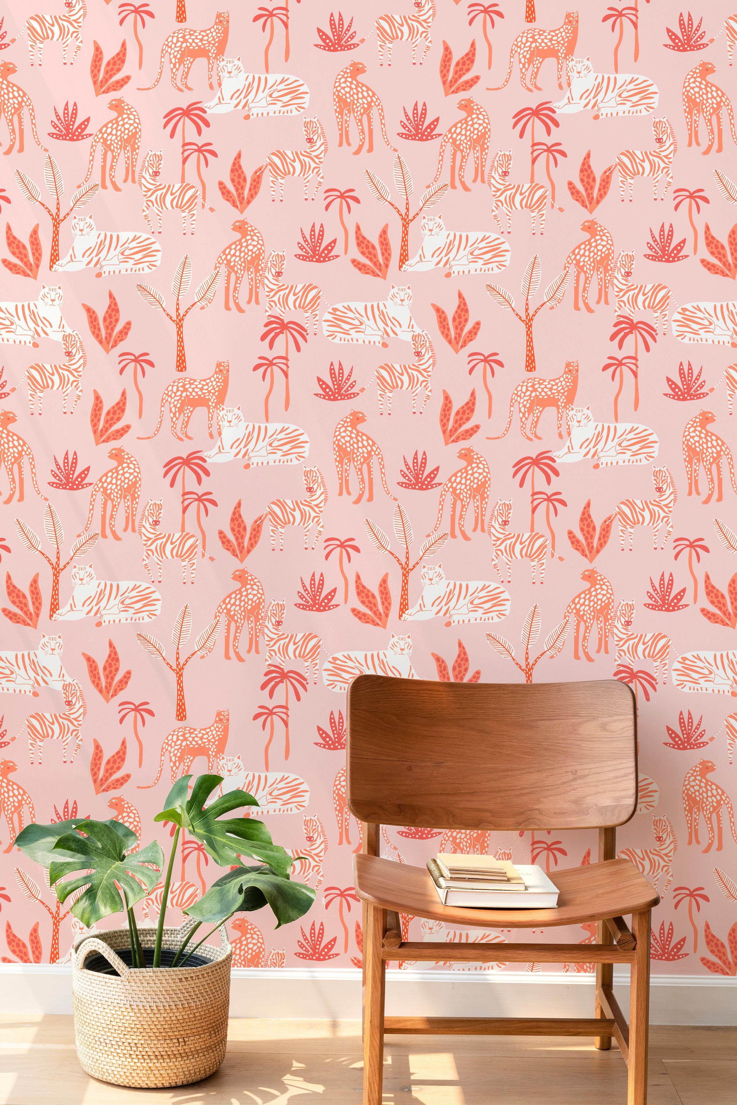A cozy reading nook decorated with Sunset Safari wallpaper, featuring colorful illustrations of safari animals and tropical plants in shades of orange, coral, and white on a pink background. The space includes a wooden chair and a potted plant.