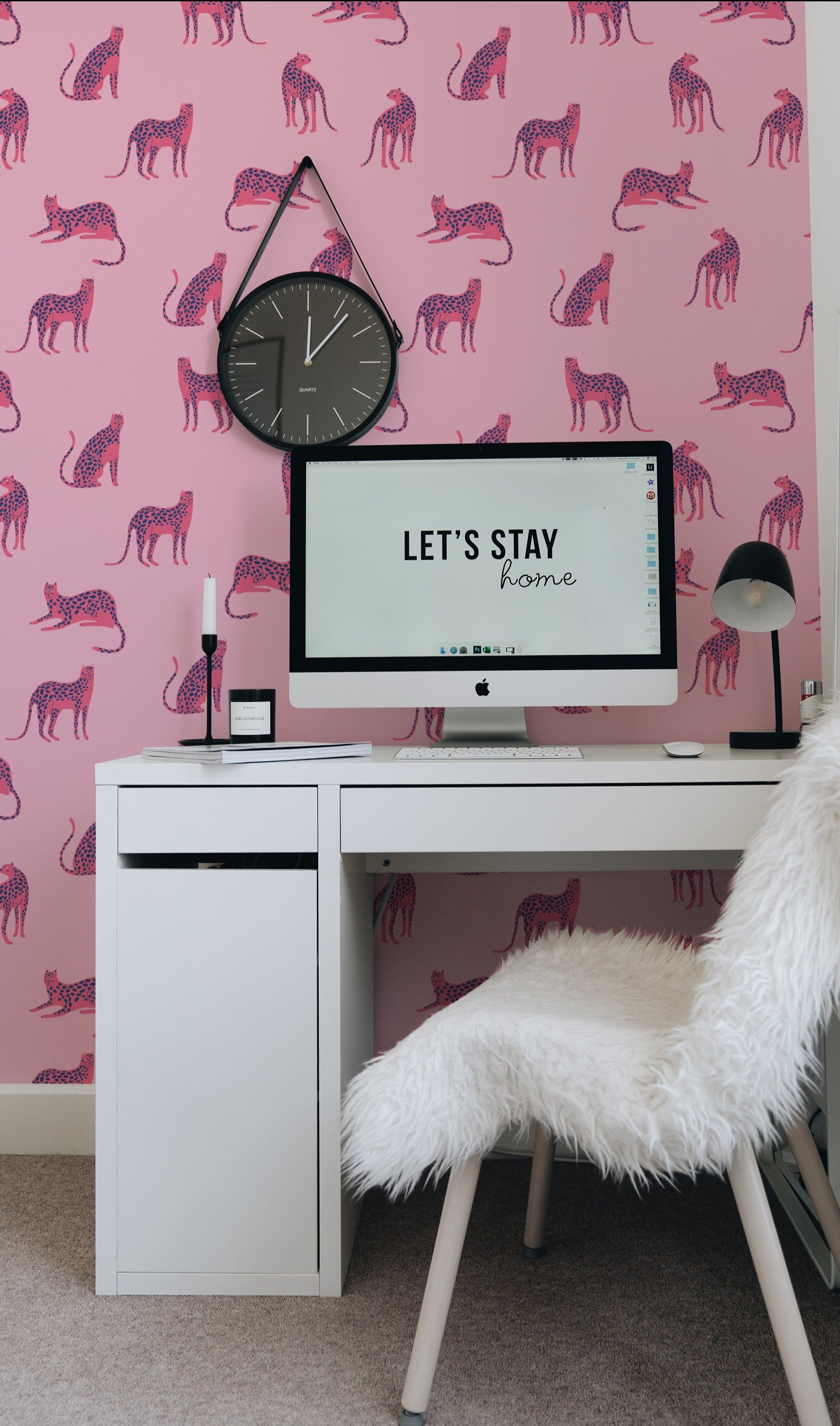 A chic home office decorated with Pink Leopard wallpaper, featuring pink and blue leopard illustrations on a pink background. The workspace includes a white desk, a fluffy chair, a clock, and a computer screen displaying 'Let's Stay Home'.