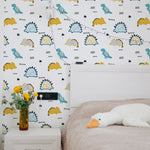 A child's bedroom showcasing the Dino Days Wallpaper as a cheerful backdrop. The room features a bed with a plush duck toy, a white bedside table with vibrant yellow flowers, and fairy lights draped across the wall, enhancing the playful theme of the wallpaper. This setup offers a lively and imaginative space, perfect for sparking creativity in young minds.