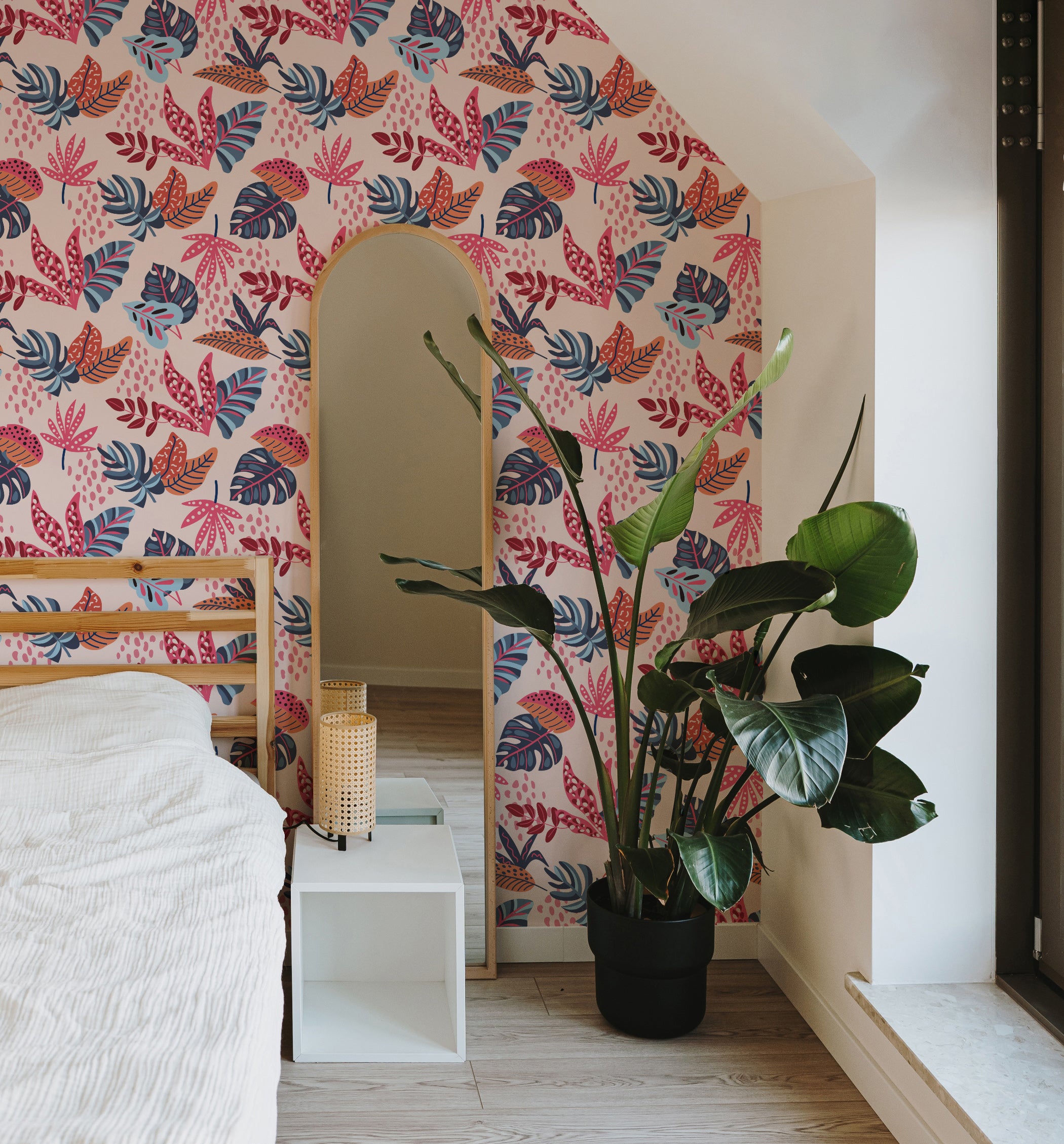 A modern bedroom decorated with Exotic Paradise wallpaper, featuring bold tropical leaves in vibrant pink, blue, and orange tones on a light beige background. The room includes a wooden bed, a tall plant, and a minimalist mirror.