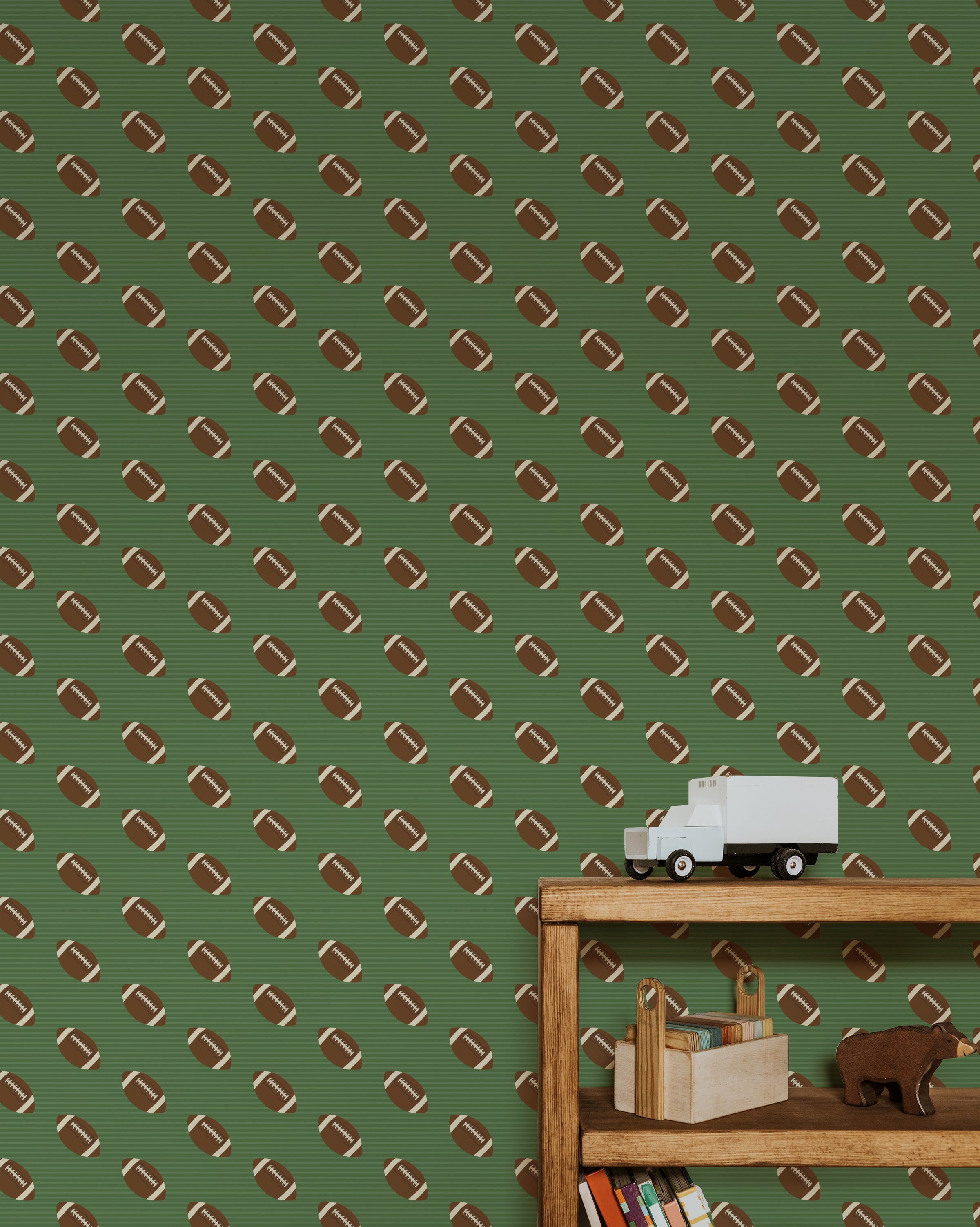 A playful children's room decorated with Football Frenzy wallpaper, showcasing a repeating pattern of brown footballs with white stripes on a green striped background. The room features a wooden bookshelf filled with toys and books.