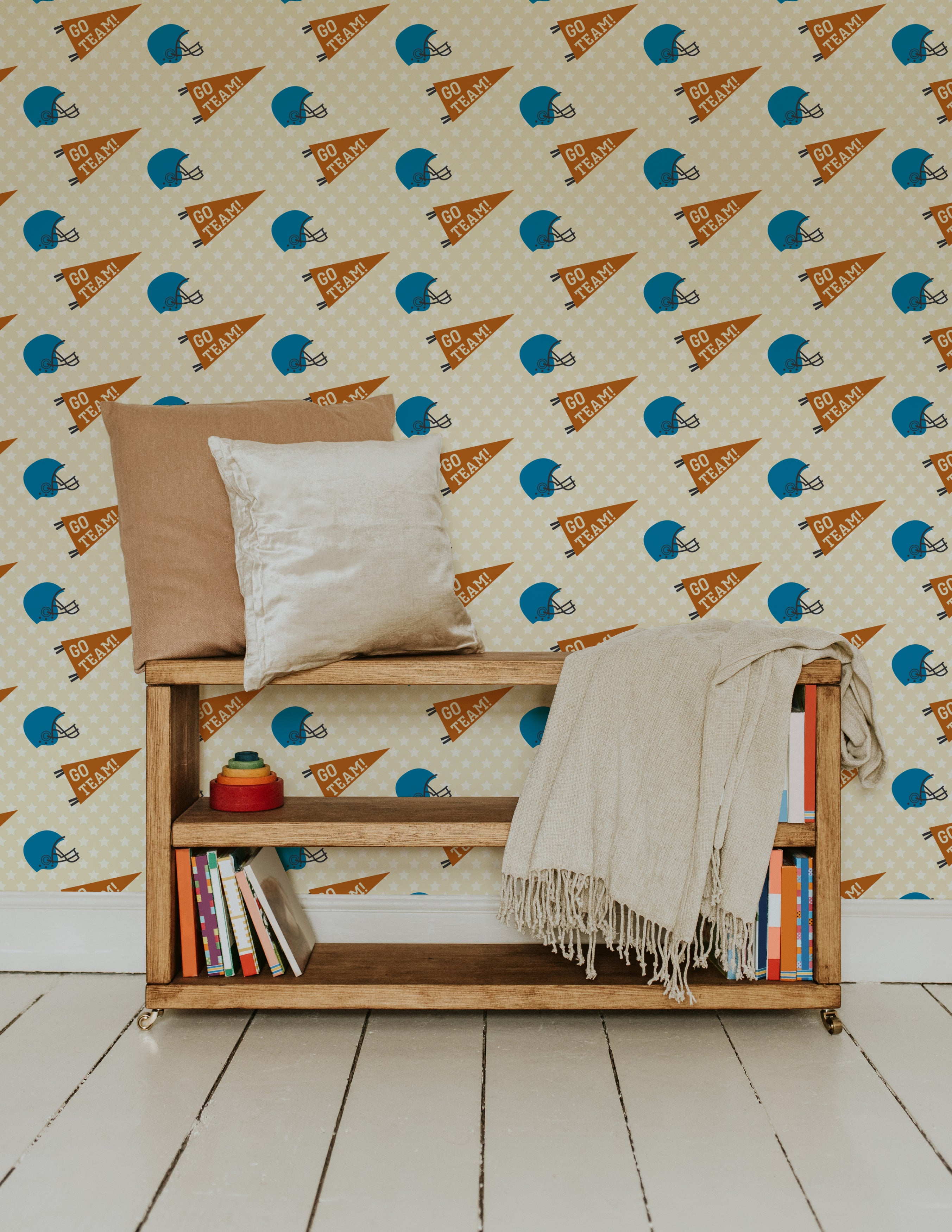 A cozy reading nook with a wooden bookshelf and cushions, decorated with Game Day wallpaper. The wallpaper features blue football helmets and orange 'Go Team!' pennants on a beige star-studded background.