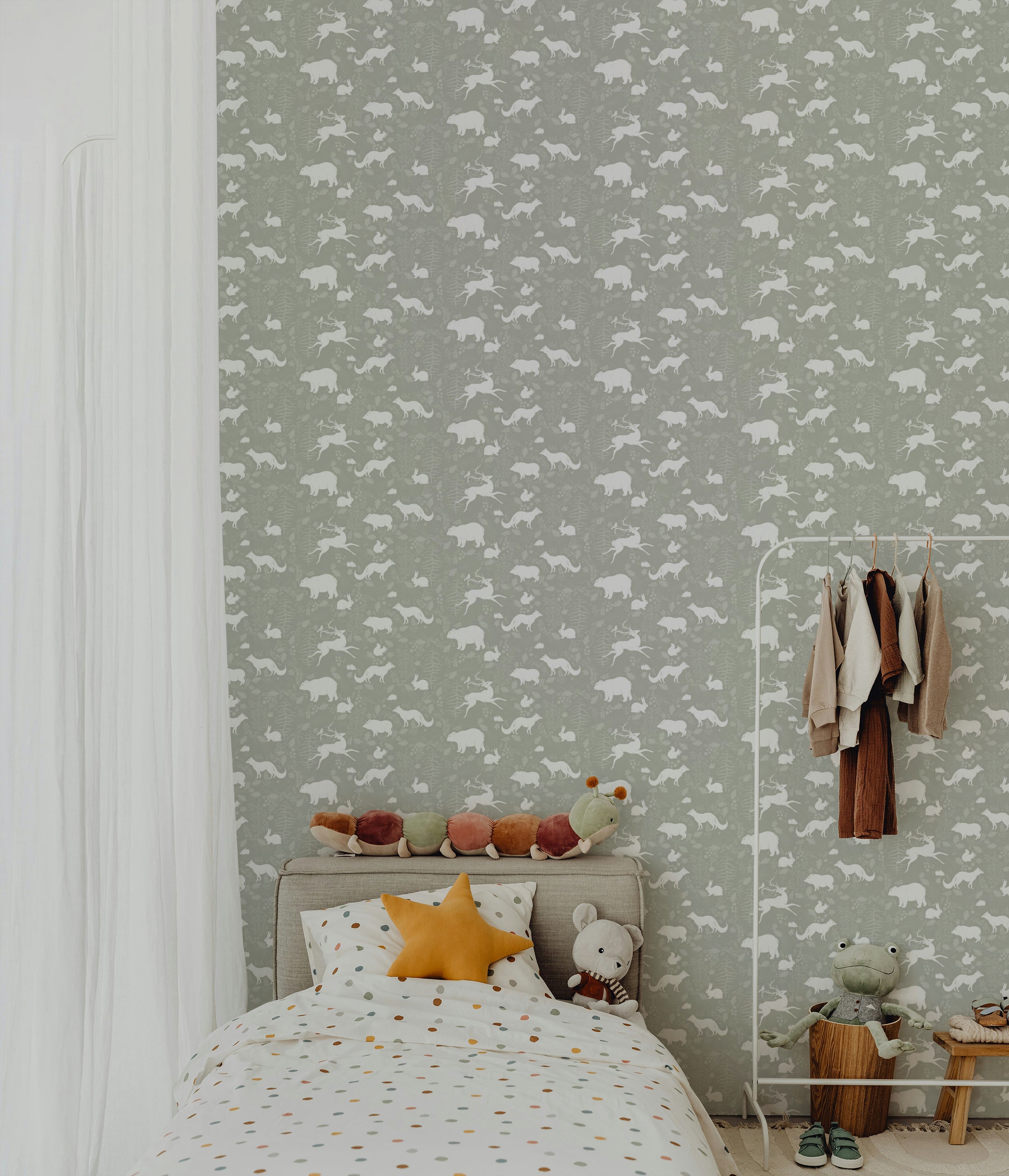 A cozy kids' room decorated with the Forest Silhouettes Wallpaper, featuring white animal silhouettes on a muted green background. A bed with colorful plush toys and a star-shaped pillow, along with a clothing rack and storage bin, create a playful and inviting space.