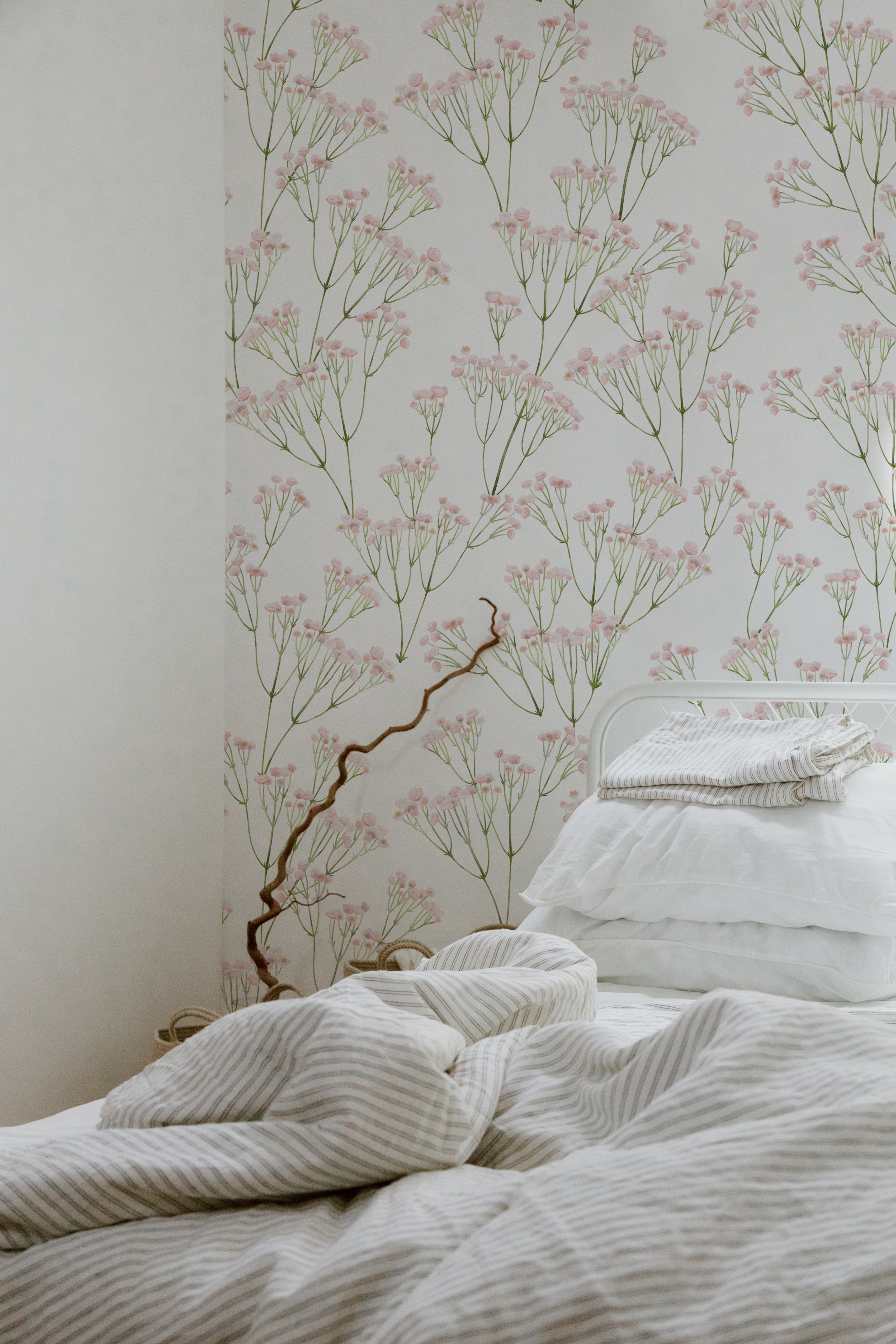 A tranquil bedroom setting displaying the Le Marchand de Fleurs Wallpaper - 75", where the wallpaper’s light pink flowers and green stems enhance the soothing atmosphere, complementing the simple white bedding and natural light.