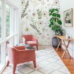 an interior scene with "Botanical Muse Wallpaper - 75"," gracing the wall of a bright, cozy sitting area. The wallpaper's lush floral pattern complements the room's natural light, enhancing the space with an organic, refreshing feel. Velvet pink armchairs and a wooden desk with a potted plant and decorative items complete this inviting nook.