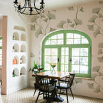 A residential dining room beautifully decorated with the Botanical Vines Wallpaper - 75". The room features a large arched window with green trim, complementing the green leaf motifs of the wallpaper. A dark wooden dining set and a classic chandelier add a touch of traditional charm, while built-in wall niches display decorative items, enhancing the room's cozy ambiance.