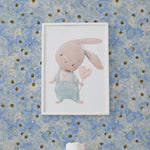 A charming nursery room decorated with the Blue Fleur Wallpaper - 25, featuring a beautiful pattern of large blue cosmos flowers with yellow centers. The wallpaper provides a vibrant backdrop to a framed illustration of a cute, pink bunny and a plush white teddy bear sitting on a white table.