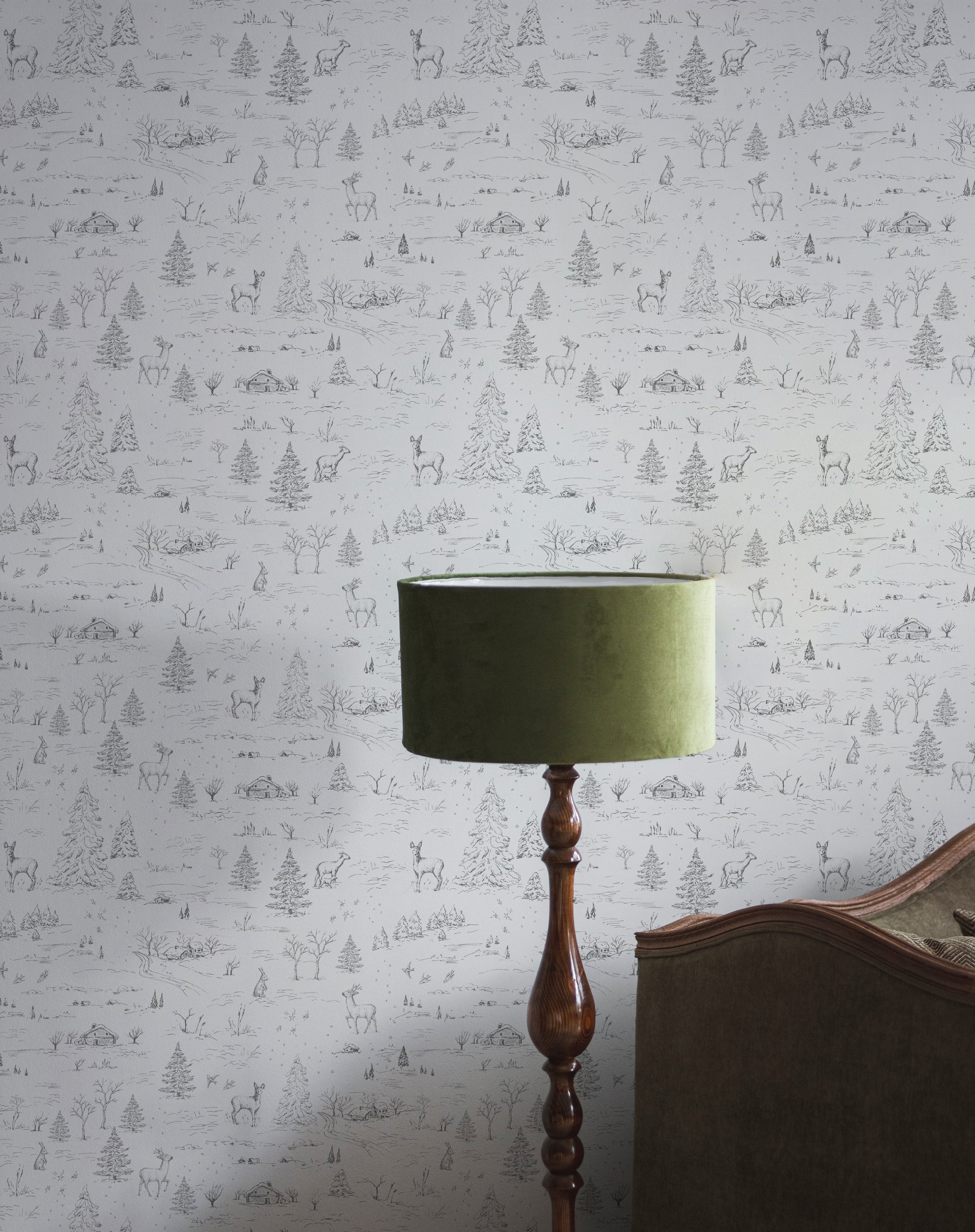 A chic room corner featuring a wall adorned with a winter wildlife sketch wallpaper, displaying intricate scenes of deer, trees, and cabins in a charming monochrome style, complemented by an elegant, vintage floor lamp with a green shade