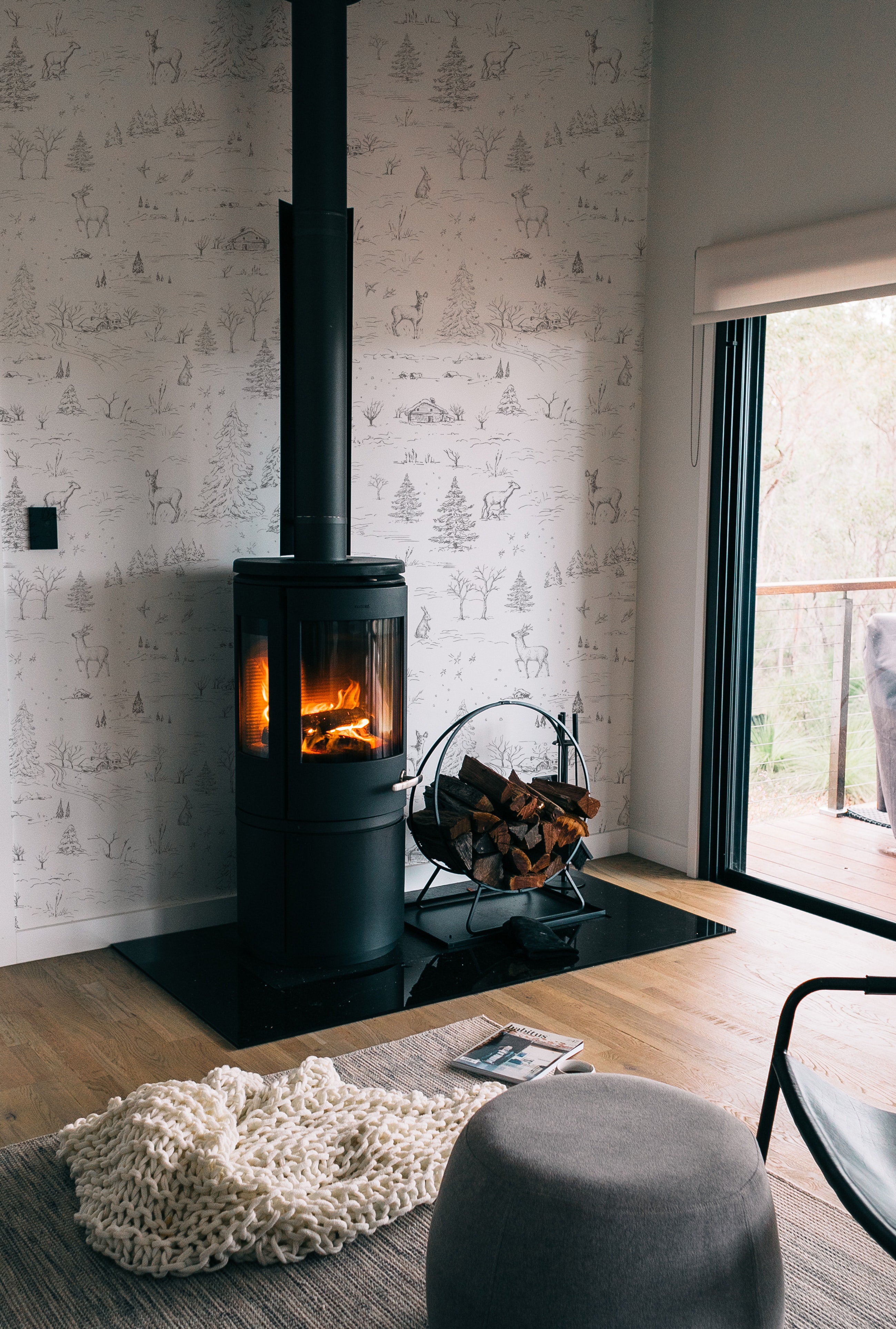 A stylish interior showcasing the winter wildlife wallpaper with its detailed sketches of forest animals and snow-covered landscapes, adding a serene and rustic charm to the room, complemented by a modern wood-burning stove.